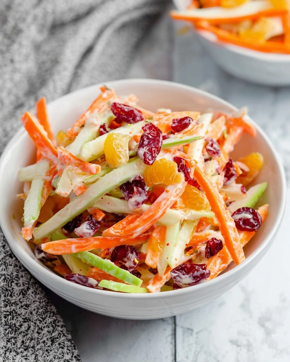 A white bowl filled with a colorful fruit and vegetable salad sits on a white marbled texture. The salad has three main layers: thin, long orange carrot strips, green apple slices with skin, and creamy white chunks of pear. Bright red dried cranberries and small orange mandarin segments are scattered throughout. The salad looks fresh and lightly coated with a creamy dressing. A grey cloth is blurred in the background, adding softness to the scene. Photo taken with an iphone --ar 4:5 --v 7