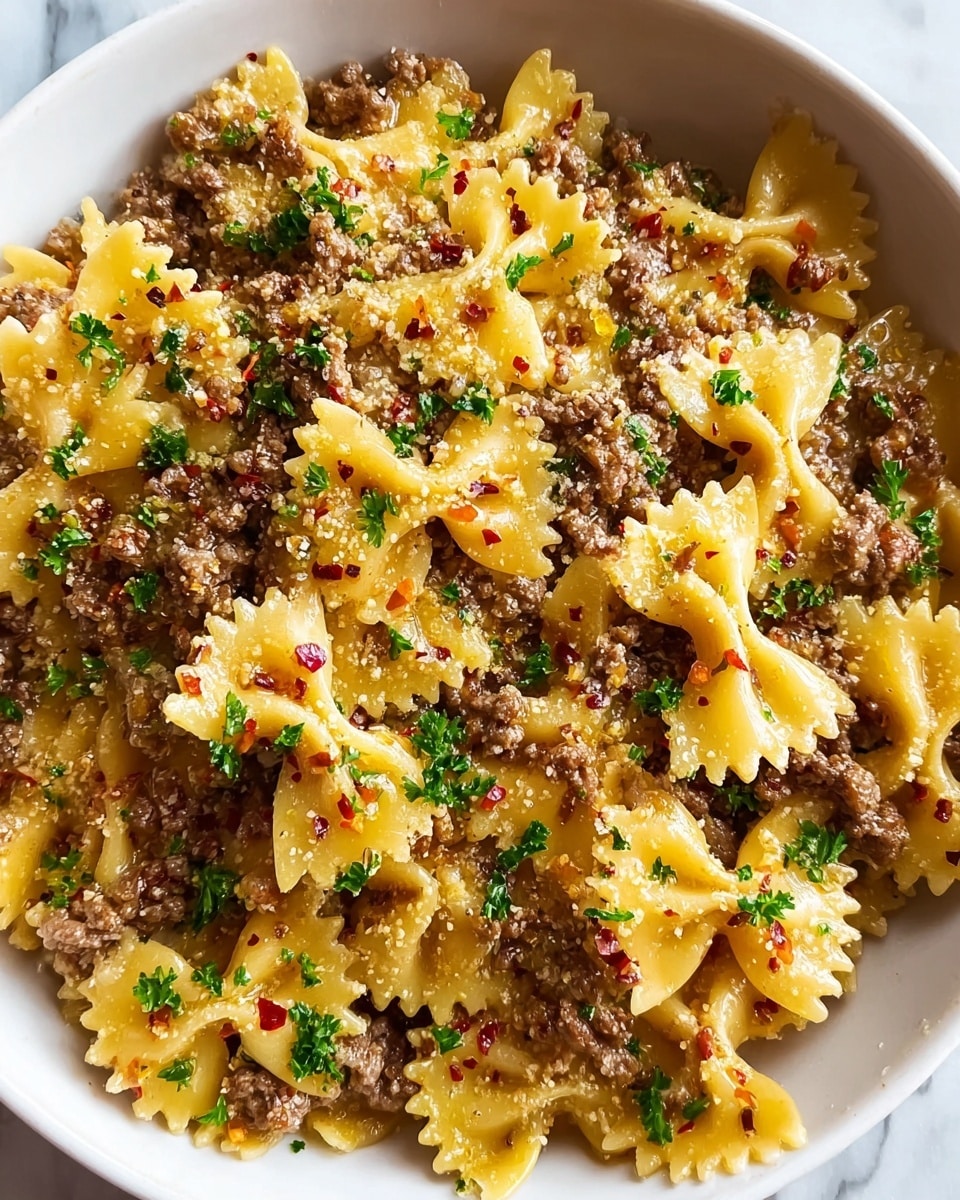 A close-up view of a white bowl filled with farfalle pasta mixed with small pieces of browned ground meat. The pasta is yellow and has a smooth, slightly shiny texture. The meat is crumbled and cooked, with a light brown color. The dish is topped with finely chopped green herbs scattered across the surface and sprinkled with grated cheese and red chili flakes, adding white and red specks. The mix looks creamy and well blended, set against a white marbled background. Photo taken with an iphone --ar 4:5 --v 7