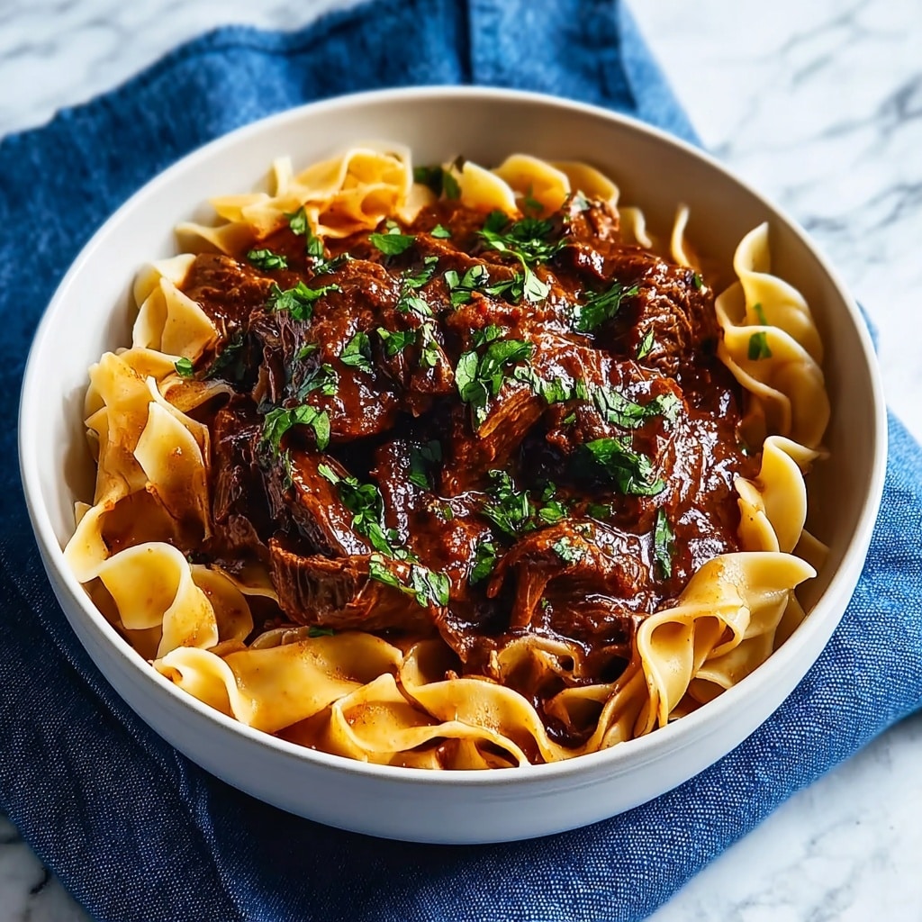 A white bowl filled with wide, flat noodles that have a smooth, slightly wavy texture and a warm golden color as the first layer. On top of the noodles is a thick layer of dark brown beef stew with visible chunks of tender meat soaked in a glossy, rich sauce. The stew layer is garnished with fresh green chopped herbs, adding a touch of brightness. The bowl is placed on a blue cloth with a white marbled surface beneath it. Photo taken with an iphone --ar 4:5 --v 7