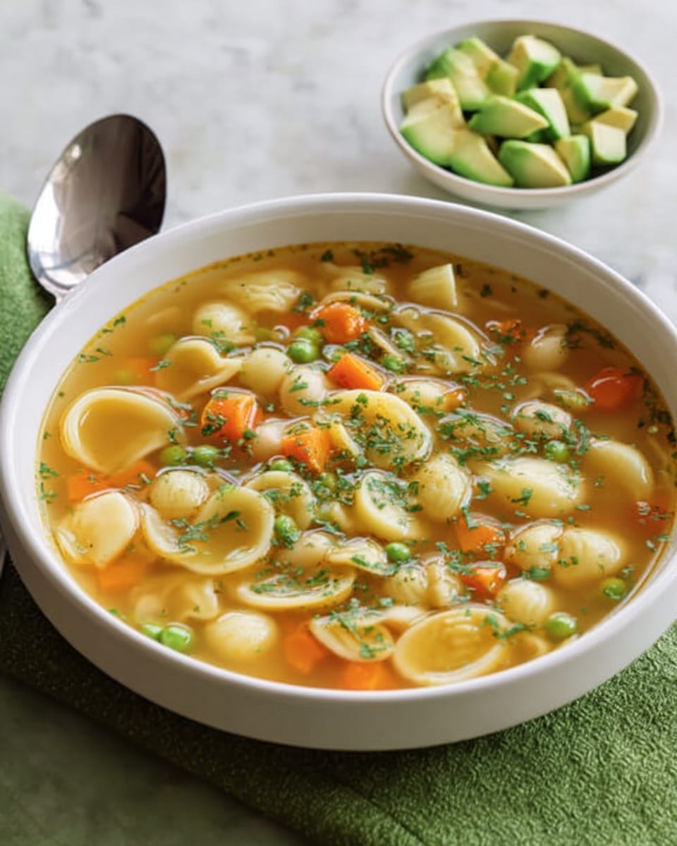 The image shows a white bowl filled with a clear soup that has several layers of ingredients. The soup contains small shell-shaped pasta that are light beige, thin slices of light yellow potatoes, small green peas, and thin strands of orange carrots. The surface of the soup is sprinkled with small bright green herb leaves, giving it a fresh look. In the back right, a small white bowl holds sliced avocado pieces that are light green. A silver spoon and a green cloth napkin sit near the bowl on a white marbled surface. Photo taken with an iphone --ar 4:5 --v 7