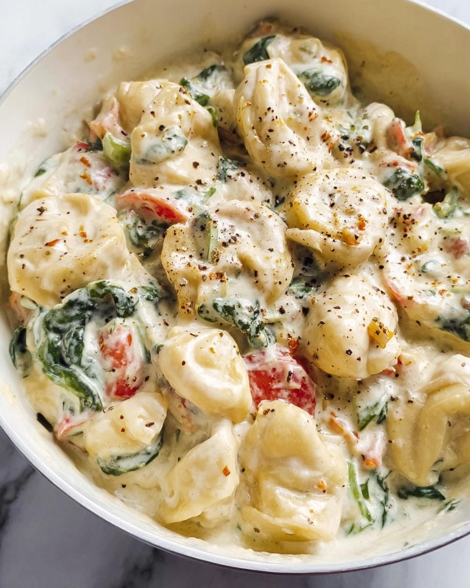 A close-up of a white plate filled with creamy tortellini pasta covered in a thick, light beige cheese sauce with small pieces of green herbs and pink bits mixed throughout; the pasta has a soft, rounded shape with some pieces folded over, and the top is sprinkled with grated white cheese, adding a crumbly texture. The plate is placed on a white marbled surface. Photo taken with an iphone --ar 4:5 --v 7