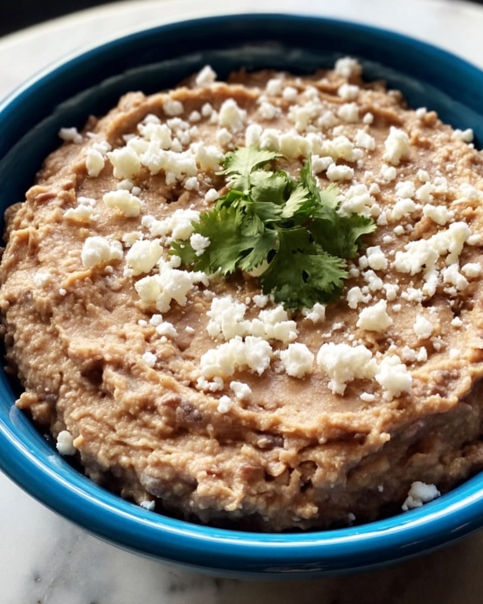 A close-up view of a creamy, smooth layer of light brown refried beans spread evenly inside a white plate, topped with small white crumbles of cheese scattered all over and a small fresh green cilantro leaf at the center for garnish, all set against a white marbled texture background. photo taken with an iphone --ar 4:5 --v 7
