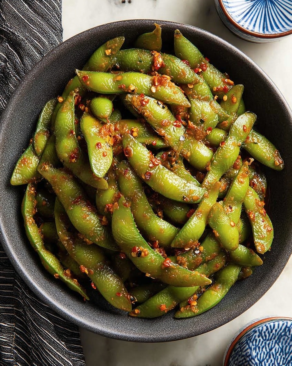 The image shows a dark gray bowl filled with cooked green edamame pods that look slightly shiny from oil. The pods have small pieces of red chili flakes, black sesame seeds, and chopped garlic scattered over them, giving a textured, spicy look. The bowl sits on a white marbled surface with a striped black-and-white cloth partly visible on the left and a partly visible blue and white ceramic container on the upper right. The overall look is rustic and appetizing, with the edamame pods arranged unevenly but fully filling the bowl. photo taken with an iphone --ar 4:5 --v 7