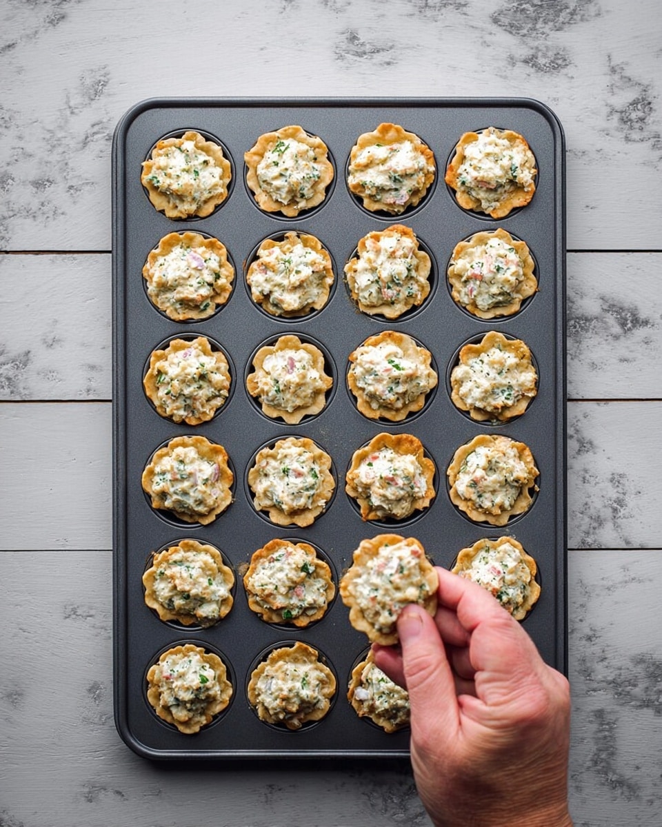 A gray baking tray filled with 31 small, round, crispy tart shells arranged closely in rows. Each tart shell is golden brown with ruffled edges, and they are filled with a creamy mixture that is white with small green herb pieces and tiny pink bits mixed in, giving it a fresh, textured look. A woman's hand reaches from the bottom center to gently pick up one of the tarts. The tray is set on a white marbled surface with subtle gray veins. photo taken with an iphone --ar 4:5 --v 7