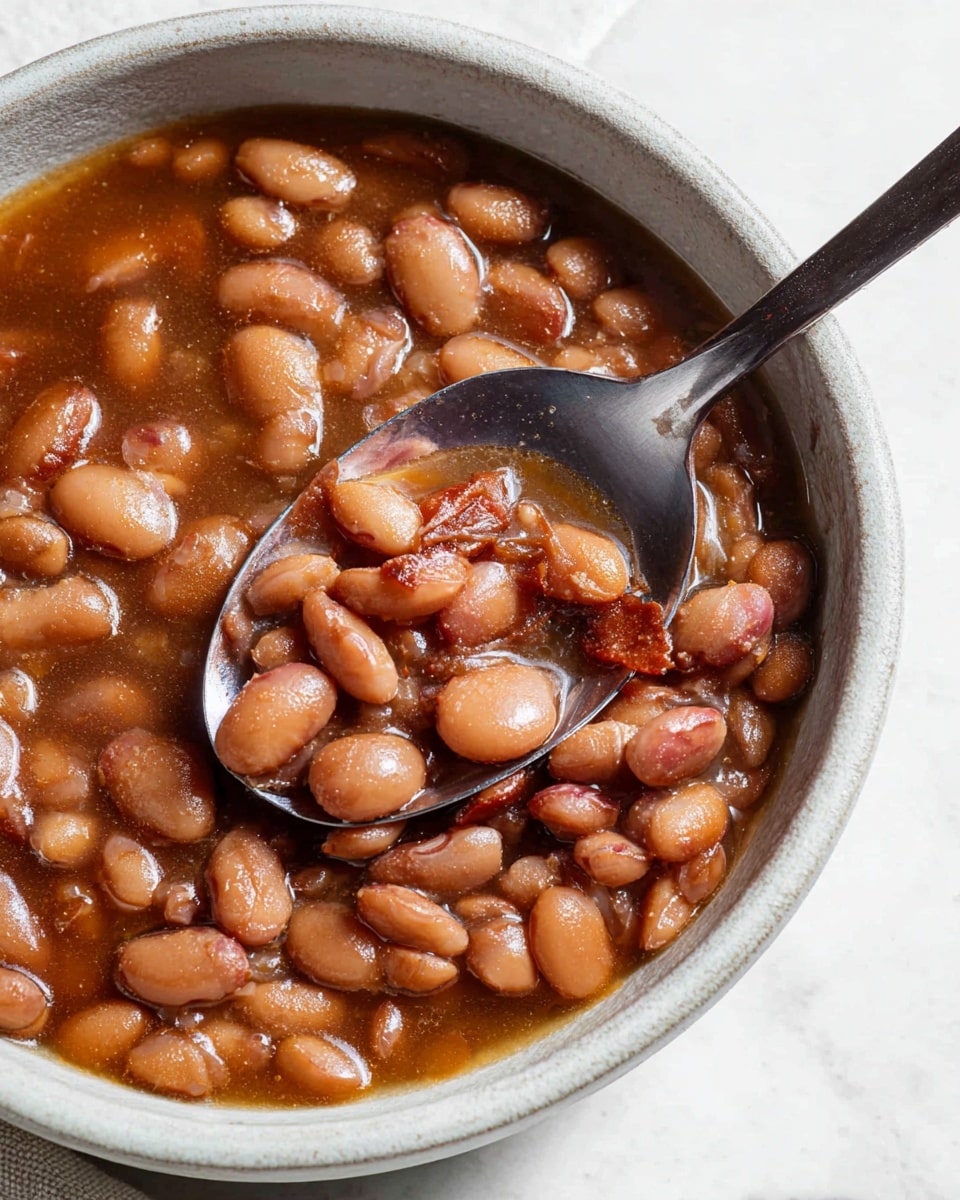 A close-up of a white ceramic bowl filled with soft cooked pinto beans in a thick brownish broth, showing a mixture of whole and slightly mashed beans with different shades of light to dark brown and reddish tones. A shiny metal spoon is partially submerged in the bowl, reflecting light and adding contrast to the warm, rich beans. The bowl sits on a white marbled surface, highlighting the natural colors and textures of the beans. photo taken with an iphone --ar 4:5 --v 7