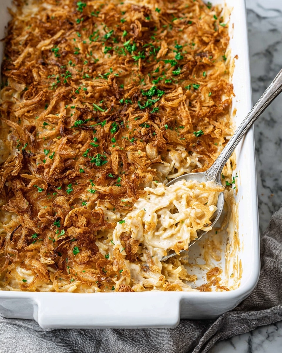 A white rectangular baking dish filled with a creamy baked casserole showing two layers. The bottom layer is creamy and pale beige with sauce and bits of noodles visible. The top layer is a thick, crispy golden-brown fried onion topping, scattered evenly with small green parsley pieces for color contrast. A silver spoon is lifting a scoop from the middle, exposing the creamy layer beneath and the crunchy texture of the topping. The dish sits on a white marbled surface with a soft, folded gray cloth on the side. Photo taken with an iphone --ar 4:5 --v 7