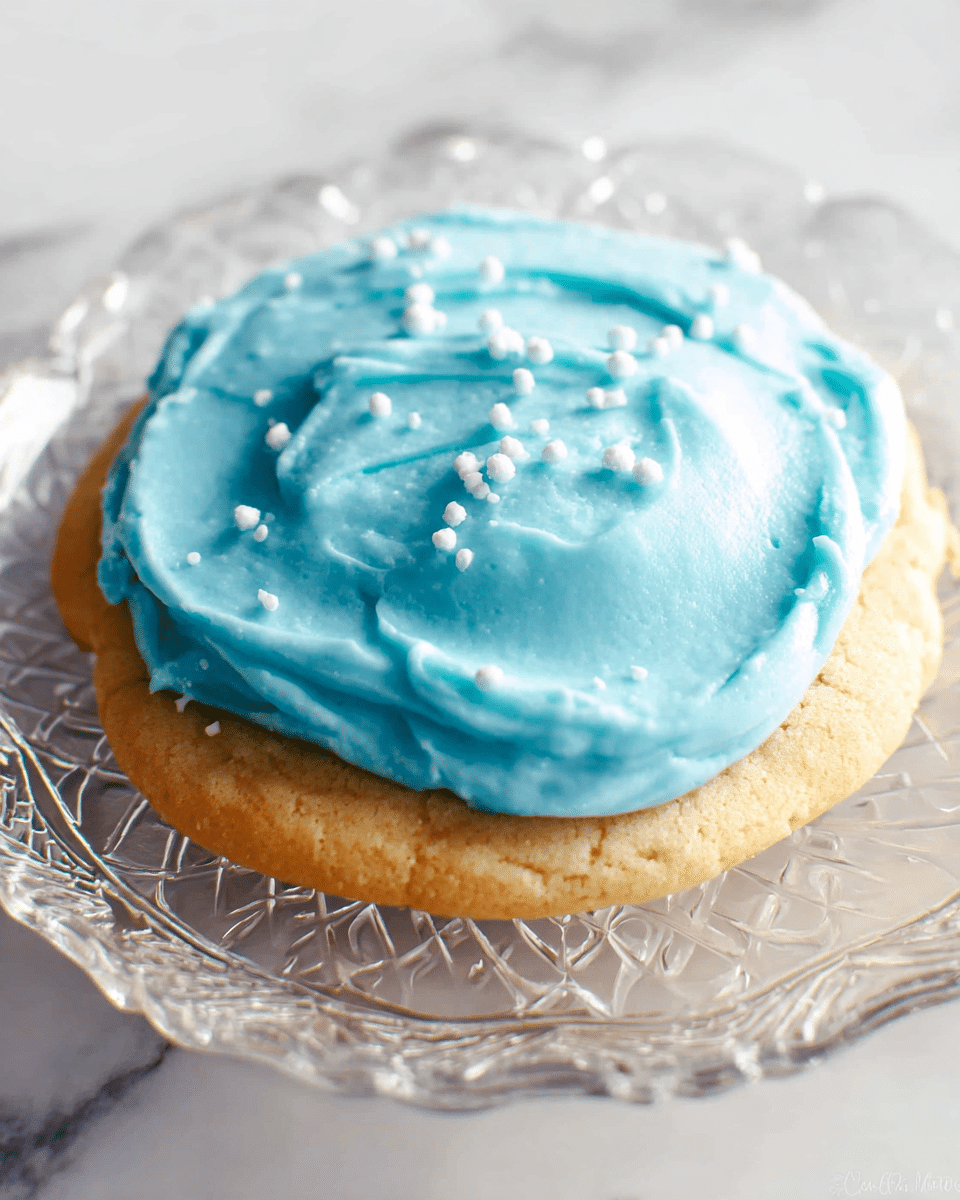 A single thick cookie with a light golden-brown color sits on a clear glass plate, which is on a white marbled texture. The cookie is topped with one smooth, thick layer of bright blue frosting that has visible spread marks and a few small white crystal-like sugar sprinkles scattered over it. The plate has an intricate pattern that adds texture to the simple presentation. photo taken with an iphone --ar 4:5 --v 7