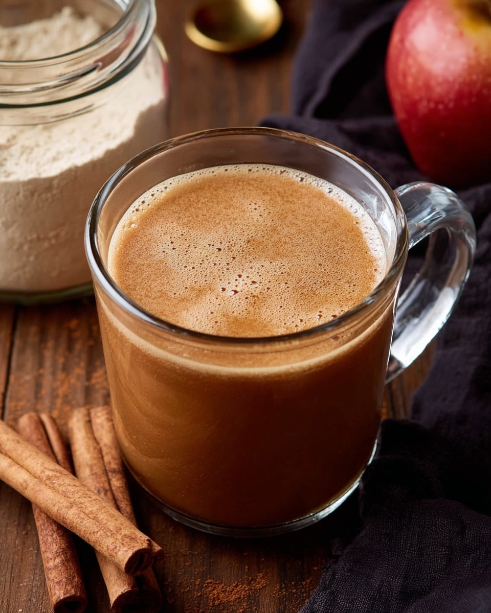 A clear glass mug filled with a smooth, warm brown drink topped with light frothy bubbles, sitting on a wooden surface with cinnamon sticks placed beside it. In the background, there is a clear jar filled with a light beige powder and a golden spoon inside, along with a red apple and a dark cloth partially visible. The overall setting is simple and cozy, showing the drink as the main focus. Photo taken with an iphone --ar 4:5 --v 7