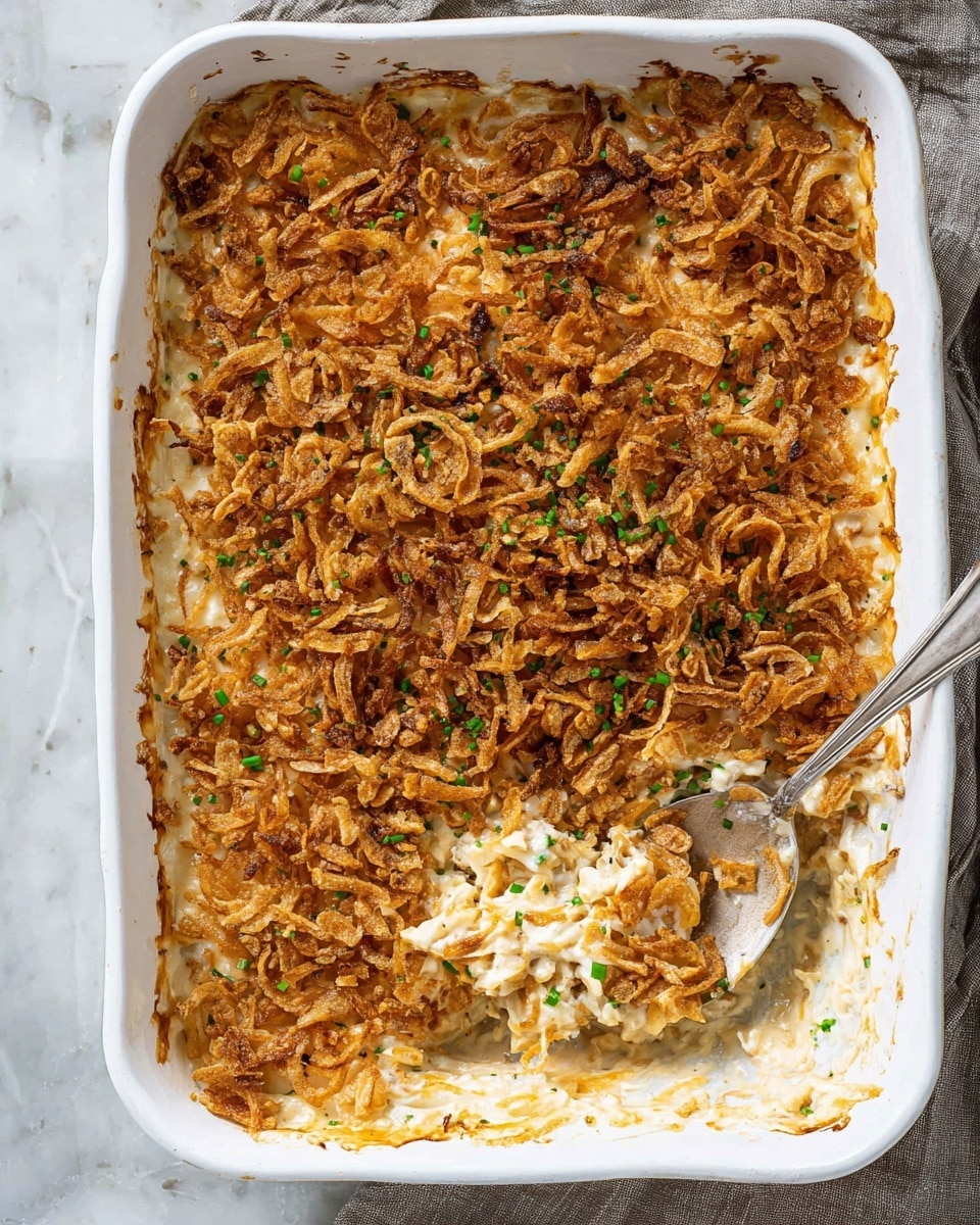 A rectangular white baking dish filled with a creamy casserole that has a golden brown, crispy topping of fried onions sprinkled with small bits of green herbs. The top layer is crunchy and uneven in texture, showing some browning at the edges. A silver spoon is scooping out a portion, revealing the creamy and cheesy middle layers beneath the crispy topping. The dish is placed on a white marbled surface with a gray cloth partially visible on the side. photo taken with an iphone --ar 4:5 --v 7