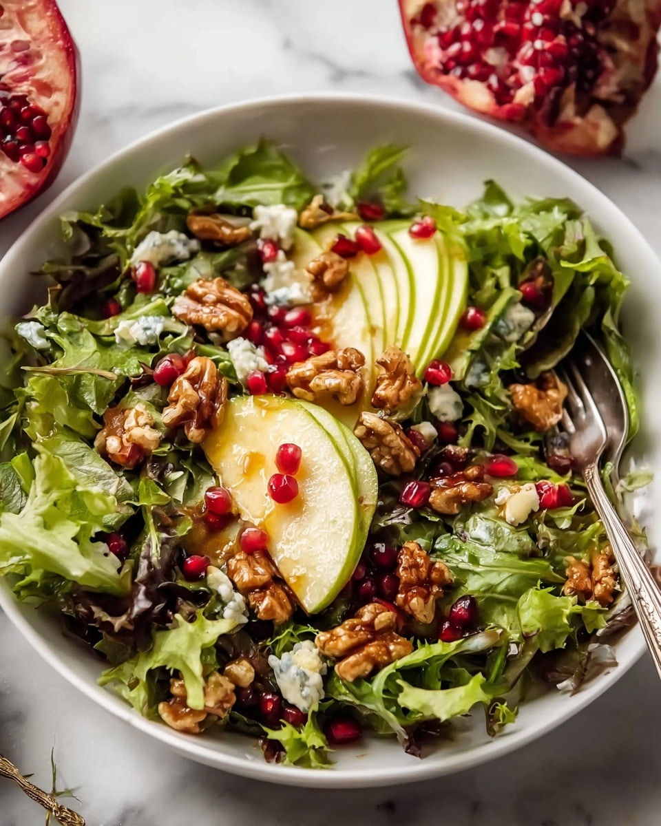 This image shows a fresh salad in a white plate on a white marbled surface. The salad has several layers starting with green mixed leafy vegetables as the base, topped with light green sliced avocado and pale yellow thin pear slices. Bright red pomegranate seeds are scattered across the salad, adding small colorful spots. Crumbled white cheese with hints of blue is spread unevenly throughout, while shiny brown walnuts are placed on top, adding texture and depth. A silver fork is resting on the side of the plate, partially in the salad. photo taken with an iphone --ar 4:5 --v 7