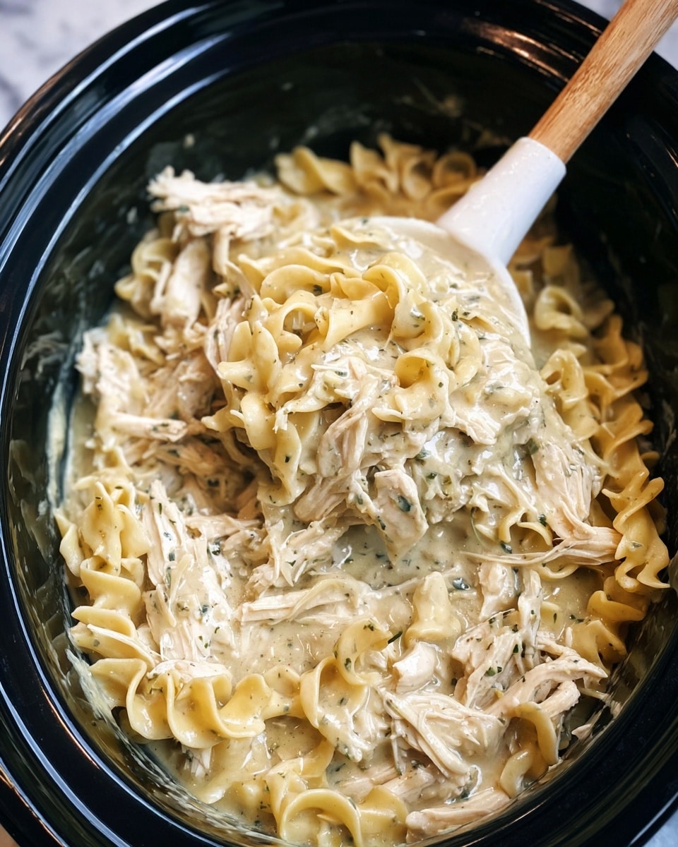 A creamy chicken noodle soup is shown inside a black slow cooker with a white spatula that has a wooden handle resting in it. The dish has two main layers: spiral egg noodles with a pale yellow color and a smooth, thick cream base that has small green herb bits scattered throughout. Tender shredded chicken pieces in a soft white tone are mixed evenly with the noodles in the creamy broth. The slow cooker is set on a white marbled textured surface. photo taken with an iphone --ar 4:5 --v 7