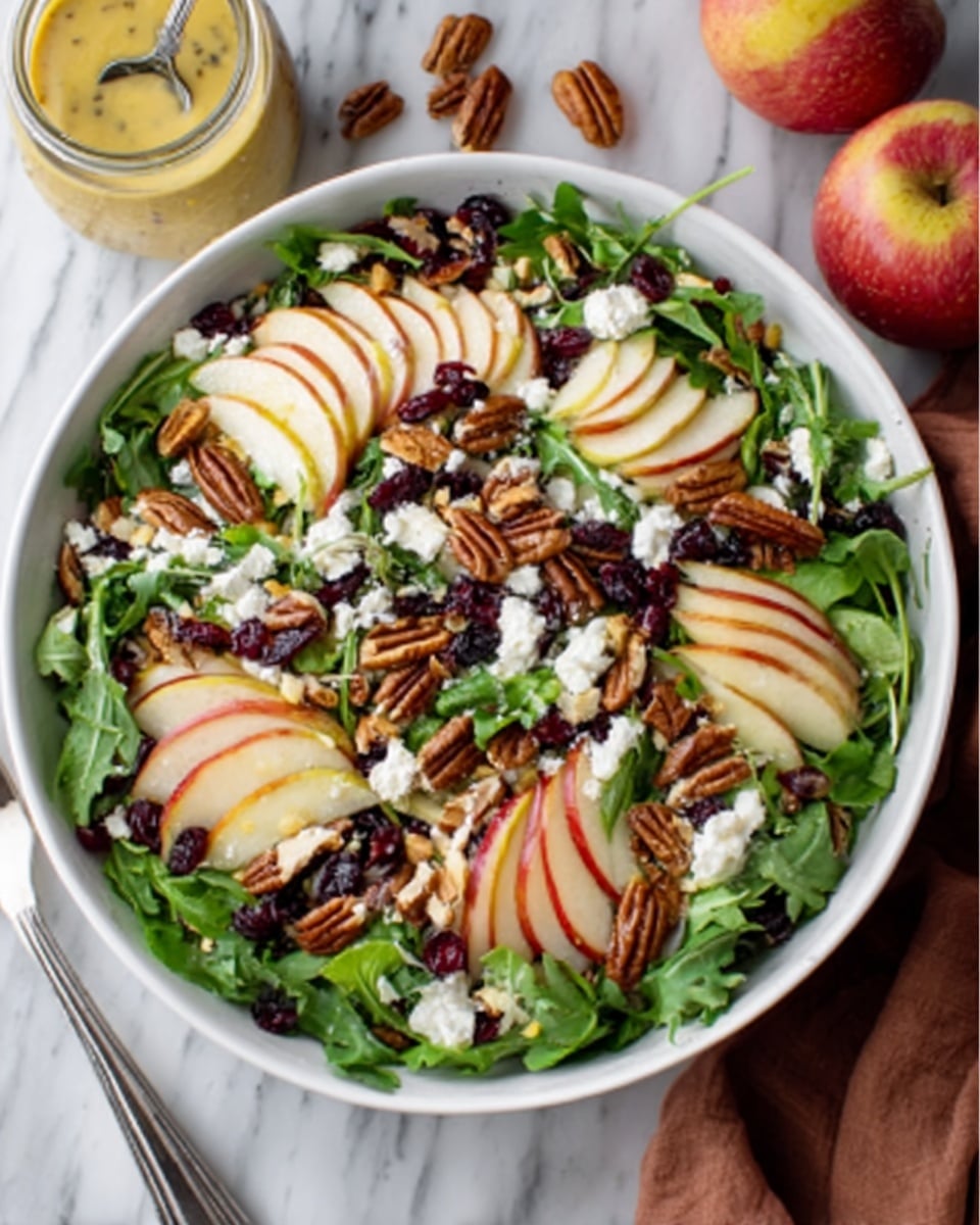A fresh salad served in a white bowl, showing a layer of green lettuce leaves at the bottom with thin slices of pale yellow apple arranged on top. Scattered across the salad are whole pecans, small white chunks of cheese, and dark red dried cranberries. The salad also has some thin slices of red onion mixed in, creating a mix of colors and textures on the fresh green bed. The background is a white marbled surface. Photo taken with an iphone --ar 4:5 --v 7