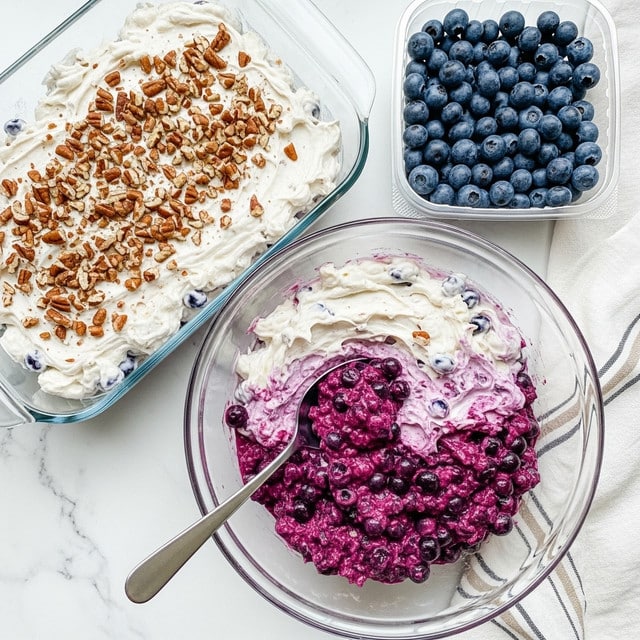 The image shows a clear glass bowl and a glass baking dish, both filled with a blueberry salad made of two visible layers. The bottom layer is a deep purple blueberry mix with a soft, chunky texture. The top layer is a creamy white mix that looks smooth and fluffy, sprinkled with small pieces of brown pecans scattered evenly. A silver spoon rests in the glass bowl, with some of the blueberry and cream mixture on it. On the right side, there is a clear plastic container filled with fresh, dark blue blueberries. All items are placed on a white marbled surface with a white and beige striped cloth nearby. Photo taken with an iphone --ar 4:5 --v 7