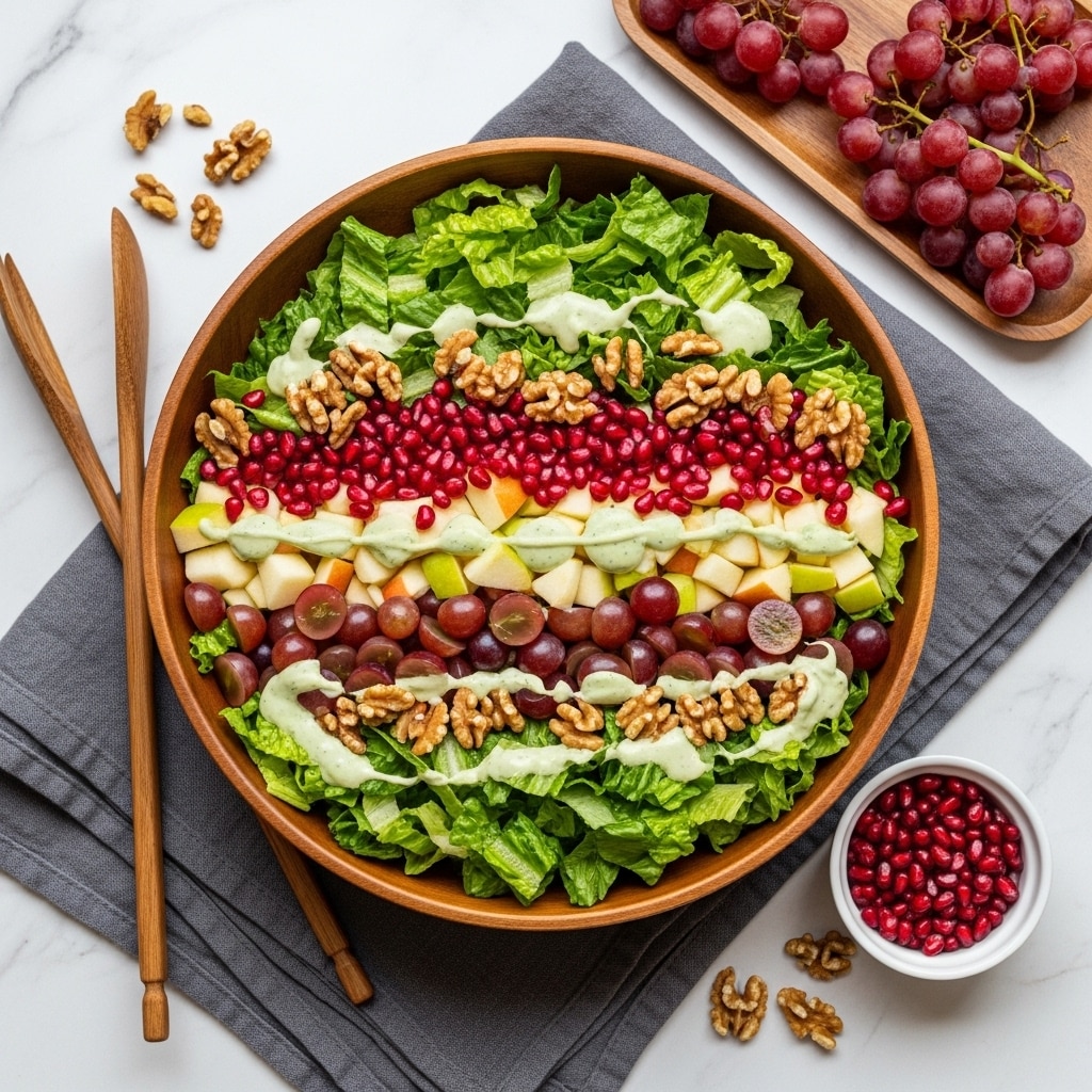 A large wooden bowl filled with a colorful salad sits on a folded gray cloth atop a white marbled surface. The salad has three main layers: a base of chopped green romaine lettuce, a middle layer of sliced red grapes and pale green apple cubes, and a top layer sprinkled with bright red pomegranate seeds and dark brown walnuts, all lightly drizzled with creamy light green dressing. Beside the bowl, there are two long wooden salad tongs, a small white bowl filled with more pomegranate seeds, clusters of red grapes on a wooden tray, and scattered walnuts and seeds adding texture to the scene. photo taken with an iphone --ar 4:5 --v 7