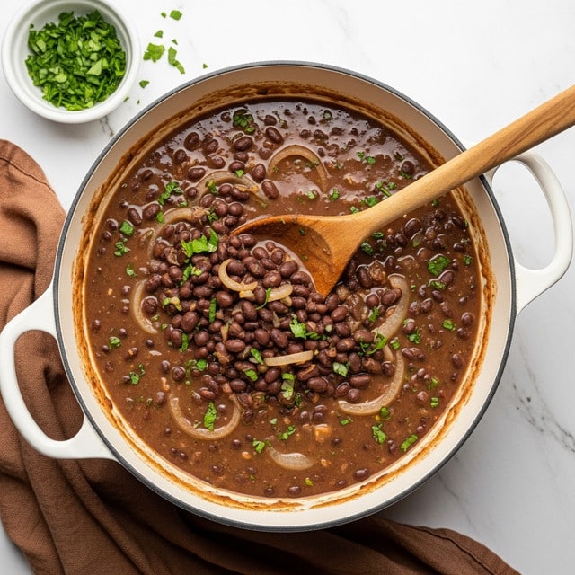 A close-up view of a large white pot filled with black bean soup, showing many whole black beans mixed with small pieces of green herbs and light-colored sliced onions, all sitting in a rich, dark brown broth. A wooden ladle with a smooth, polished texture is resting inside the pot, partially submerged in the soup, capturing the thick and chunky mixture. The pot is placed on a white marbled surface, and the overall scene looks warm and hearty. photo taken with an iphone --ar 4:5 --v 7