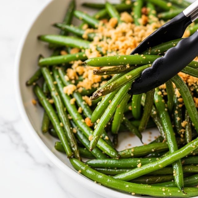 A close-up image of a white dish filled with bright green cooked green beans, slightly blistered and tossed with light golden crispy breadcrumbs scattered unevenly on top and around the beans. Black tongs are picking up a small bundle of the green beans, showing their shiny texture from a light coating of oil. The background surface is a white marbled texture. photo taken with an iphone --ar 4:5 --v 7