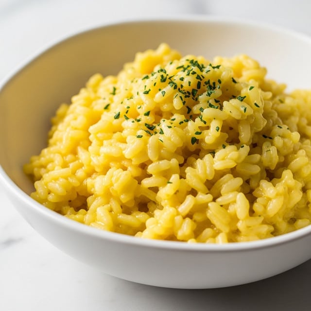 A close-up image of creamy yellow rice with a slightly shiny texture topped with small green herb specks evenly spread on top. The rice is in a white scalloped bowl placed on a white plate. The whole setup is on a white marbled background, and the dish looks soft and moist with a rich, smooth consistency. Photo taken with an iphone --ar 4:5 --v 7