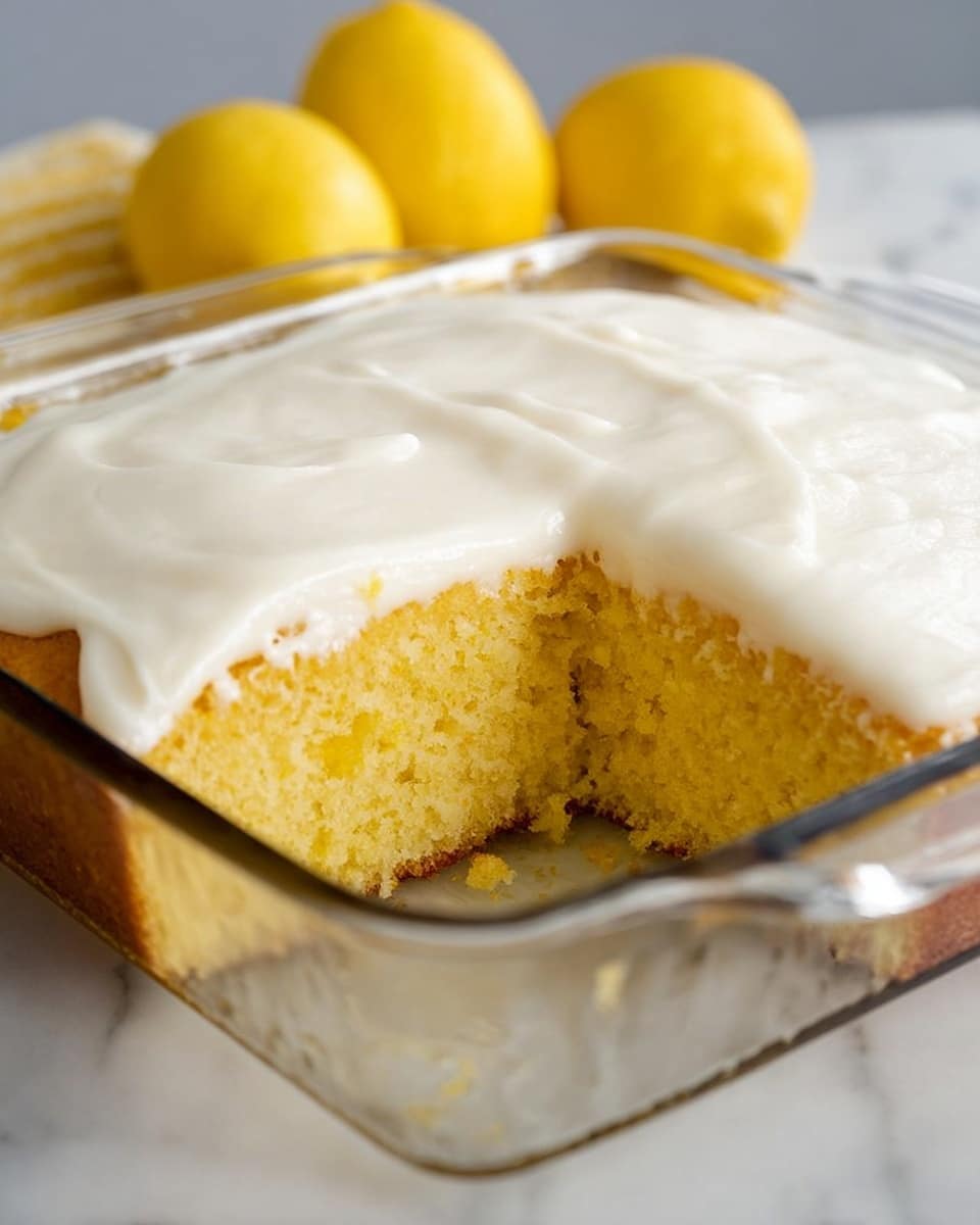 A square glass dish holds a yellow cake with a soft, moist texture, cut to show its airy middle layer. The cake is topped with a thick, smooth layer of white frosting that covers the entire top surface evenly. Behind the dish, there are two lemons that add a fresh touch to the setting, which rests on a white marbled surface. photo taken with an iphone --ar 4:5 --v 7