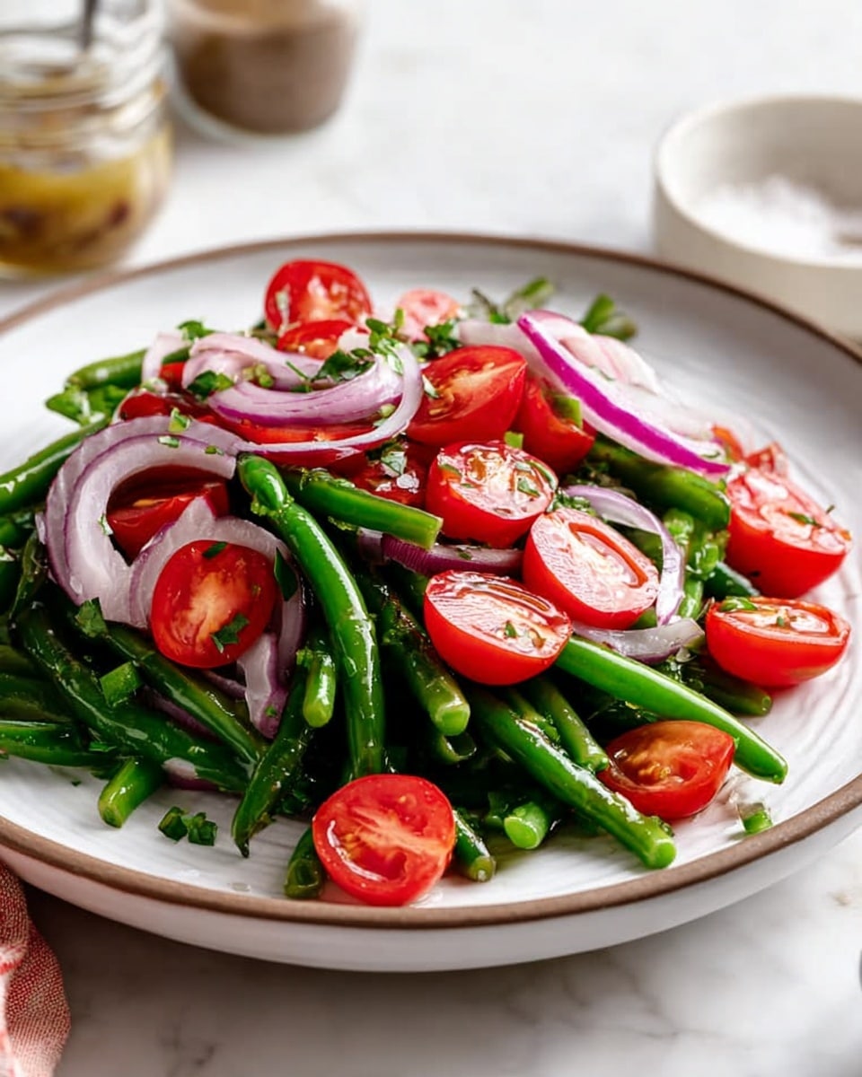 The dish is a fresh salad served on a white plate with a slightly rounded edge and a subtle brown rim. The base layer consists of vibrant green long beans that are slightly glossy, arranged in a loose pile. On top of the green beans are halved bright red cherry tomatoes, adding a juicy, smooth texture and bold color. Thin, slightly translucent slices of purple-red onions are scattered evenly across the salad, providing a light crunchy layer. Small chopped green herbs are sprinkled throughout, adding a fresh, leafy contrast. The background features a white marbled surface, with a blurred jar of dressing and a white bowl with salt and pepper just behind the plate. photo taken with an iphone --ar 4:5 --v 7