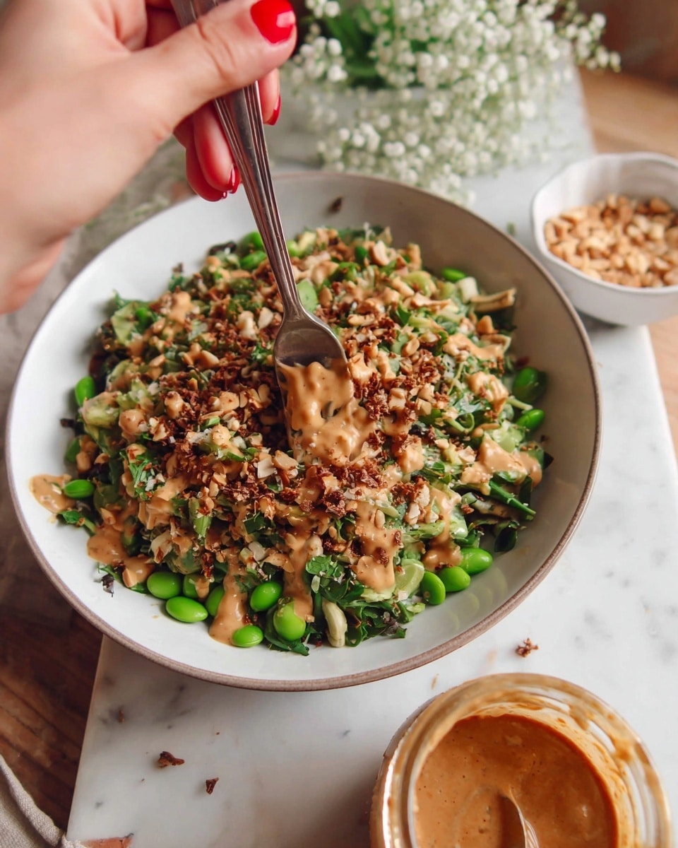 The image shows a bowl of mixed salad with several layers, starting with a base of green leafy vegetables and edamame beans, creating a fresh green layer. On top, there is a mixture of finely chopped brown and beige nuts and seeds, adding a crunchy texture. The dish is drizzled with a light brown creamy sauce that pools a bit around the top. The bowl is white, set on a white marbled surface. A woman's hand with red nail polish is holding a fork that is stirring into the salad, and in the foreground, there is a jar with more of the same creamy sauce on a spoon. Nearby, a small white bowl contains a portion of chopped nuts or dressing, and some white flowers are slightly visible to the side. Photo taken with an iphone --ar 4:5 --v 7