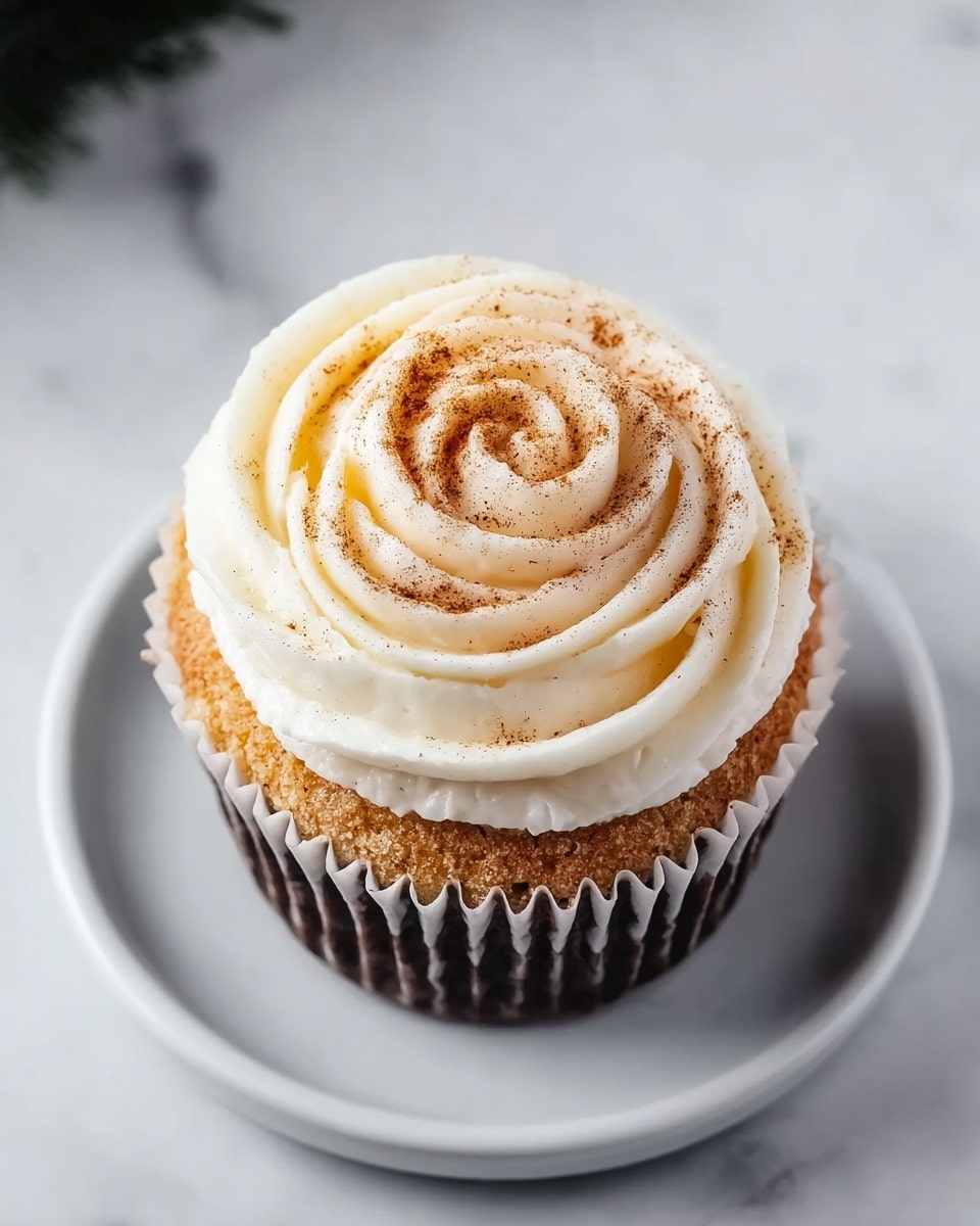 A close-up view of a single cupcake centered on a small black plate, sitting on a white marbled surface. The cupcake has two main layers: the bottom layer is a golden-brown cake with a soft texture, and the top layer is cream-colored frosting swirled in a rose shape with smooth, thick bands. The frosting is lightly dusted with a fine sprinkle of brown cinnamon powder, adding a subtle speckled effect. Around the edges, a white paper liner holds the cupcake, creating a neat border between the cake and the black plate. Around the main cupcake, partial views of similar cupcakes rest on the same white marbled surface. photo taken with an iphone --ar 4:5 --v 7