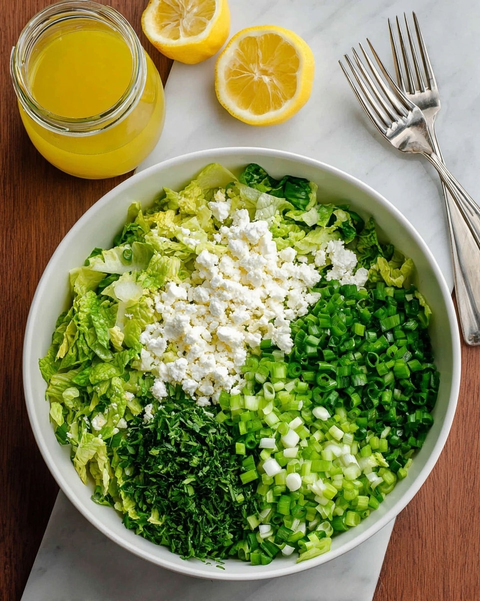 A large white bowl filled with a fresh green salad made of chopped leafy greens in different shades of green, mixed with small white crumbles of cheese scattered evenly on top. The salad has a light, moist texture that shows a mix of finely shredded and chopped leaves with small sprigs of fresh herbs adding detail. Two silver forks rest inside the bowl slightly embedded in the salad. To the right of the bowl, there are two whole bright yellow lemons set against a white marbled surface. photo taken with an iphone --ar 4:5 --v 7