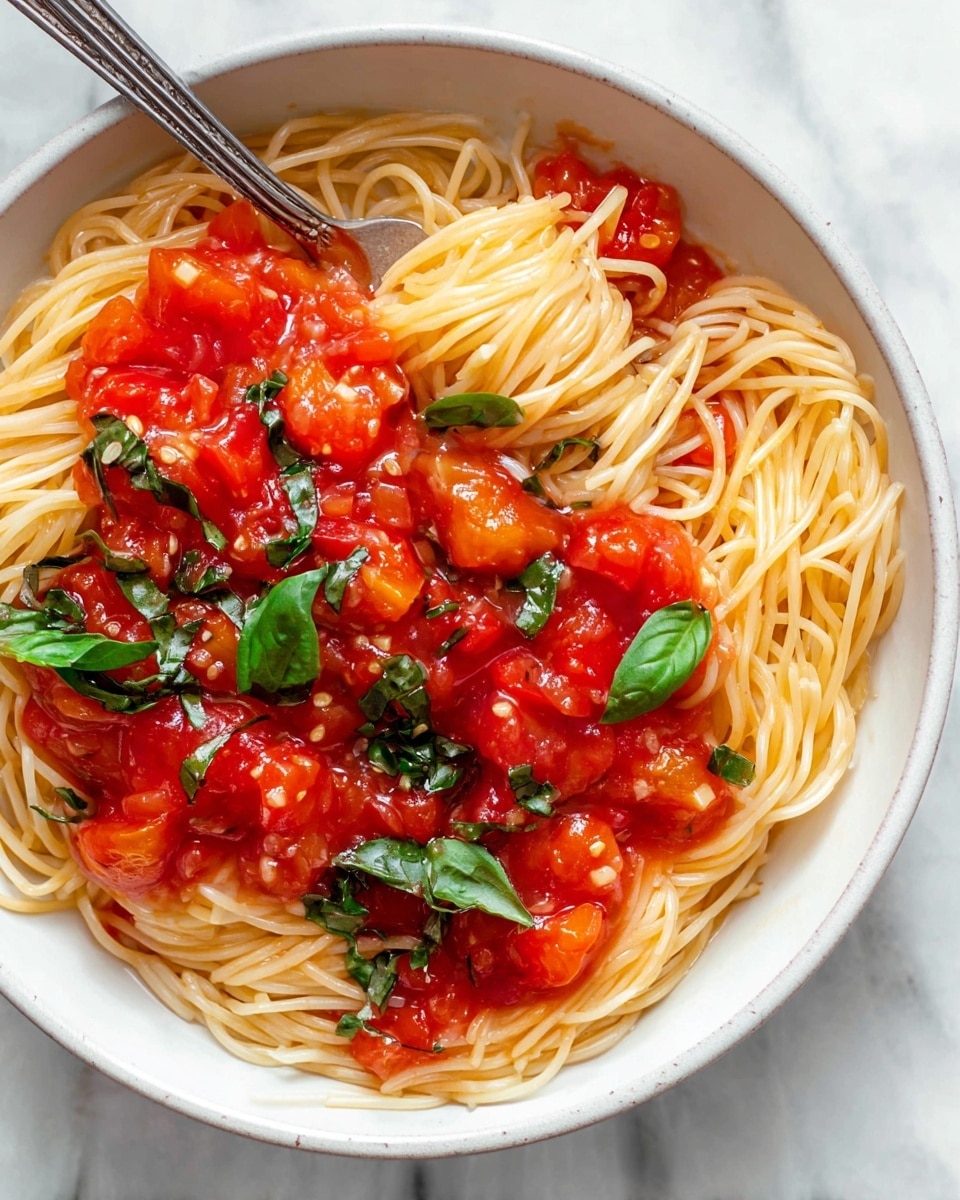 A white speckled bowl holds a simple pasta dish with two forks resting inside. The base layer is thin, pale yellow spaghetti piled loosely in the bowl. Scattered throughout and on top are bright red tomato sauce chunks with visible bits of garlic, giving a glossy texture. Fresh green basil leaves are woven within the pasta and placed on top, adding a pop of vibrant color. The bowl sits on a white marbled surface with some green basil leaves nearby and a light-colored cloth to the side. photo taken with an iphone --ar 4:5 --v 7