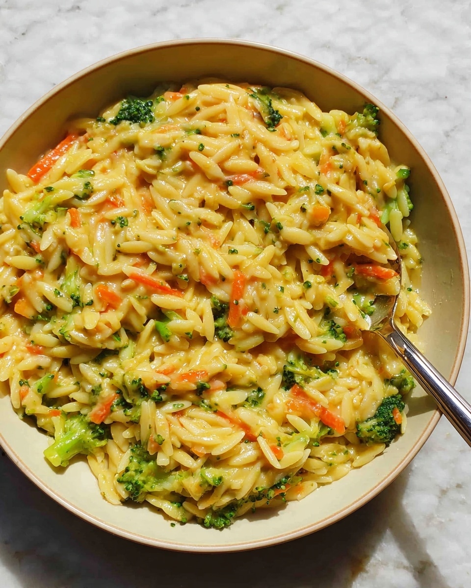 A white bowl filled with a creamy pasta dish that has three main visible layers: small pasta pieces coated in a thick yellow cheese sauce, mixed with bright green broccoli florets and small orange carrot strips. The pasta looks soft and slightly glossy from the sauce. Some pieces of the broccoli and carrot are spread evenly throughout, adding spots of color in the yellow pasta mixture. A metal spoon is partially placed on the right side of the bowl. The bowl is resting on a white marbled surface. photo taken with an iphone --ar 4:5 --v 7