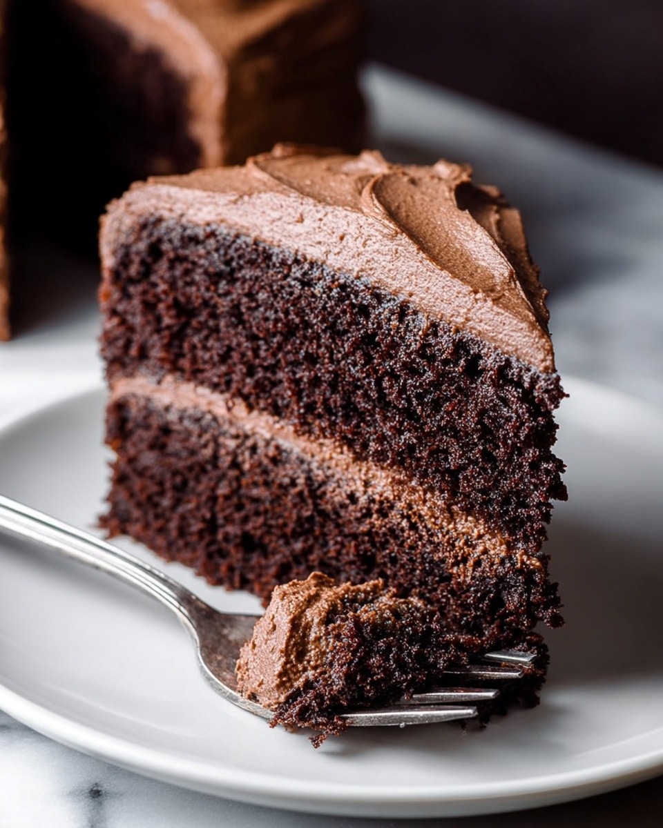 A two-layer chocolate cake is covered fully in smooth, thick chocolate frosting with visible swirls and a slightly textured surface on the top and sides, sitting on a white cake stand with a beaded edge, placed on a white marbled surface. photo taken with an iphone --ar 4:5 --v 7