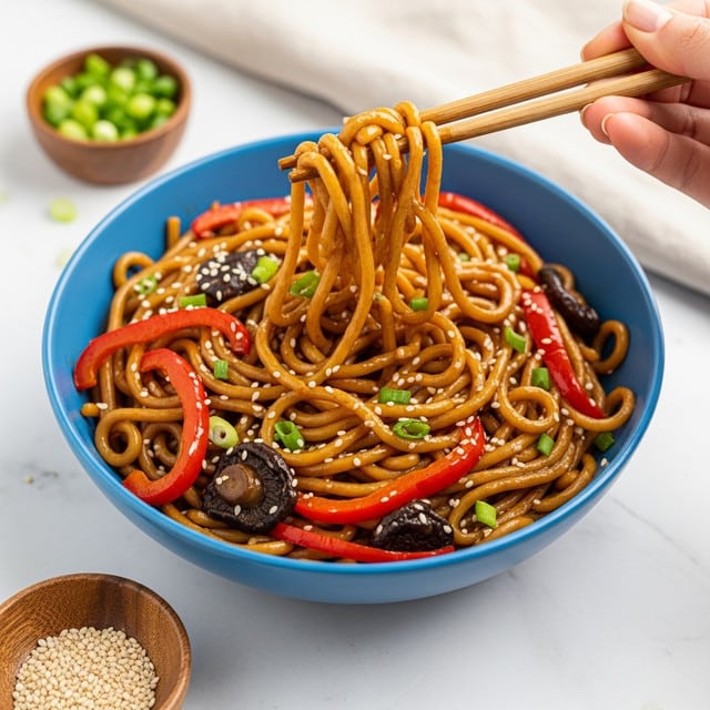 A blue bowl filled with thick, glossy brown noodles mixed with sliced red bell peppers, dark mushrooms, and small green onion pieces, all coated in a shiny, rich sauce. A woman's hand is using wooden chopsticks to lift some noodles from the bowl, showing their smooth and slightly twisted texture. The bowl sits on a white marbled surface with small wooden bowls nearby holding green onions and sesame seeds, adding a natural touch to the scene. Photo taken with an iphone --ar 4:5 --v 7