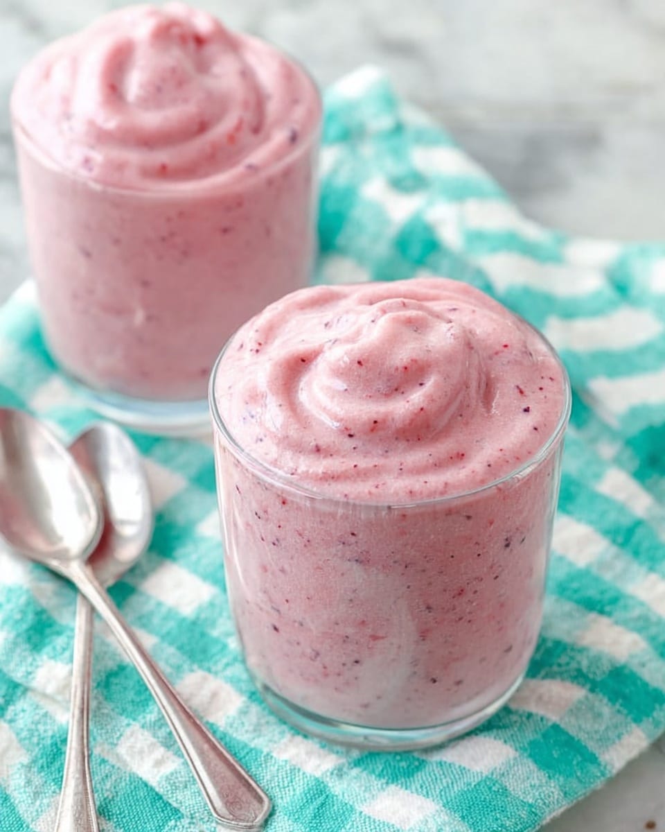 Two clear glasses filled to the top with a creamy pink smoothie that has tiny darker specks throughout, giving it a textured look. The smooth surface on each drink is softly swirled, showing a thick and airy consistency. They sit on a white marbled surface covered partly by a turquoise and white checkered cloth. Next to the glasses, there are two long silver spoons crossed over each other, adding a touch of shine. photo taken with an iphone --ar 4:5 --v 7