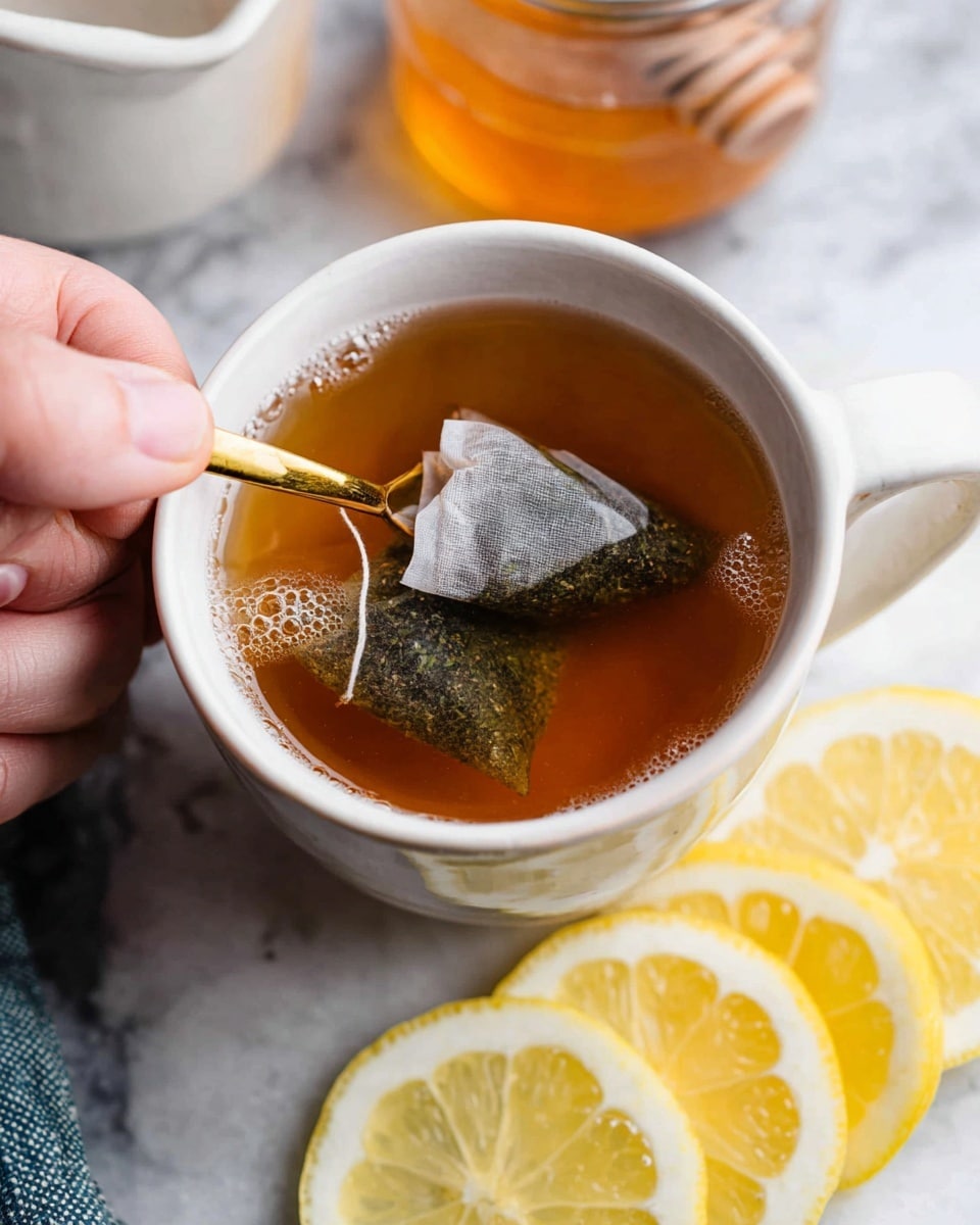 A white cup filled with dark amber tea, showing two tea bags steeping inside with visible dry herbs inside the bags, some floating on the surface; a golden spoon rests inside the cup. Behind the cup is a white container holding dark brown honey with a wooden honey dipper, a whole yellow lemon, and to the side are three yellow lemon slices bright on a white marbled surface. The foreground includes a corner of a black and white striped cloth. Photo taken with an iphone --ar 4:5 --v 7
