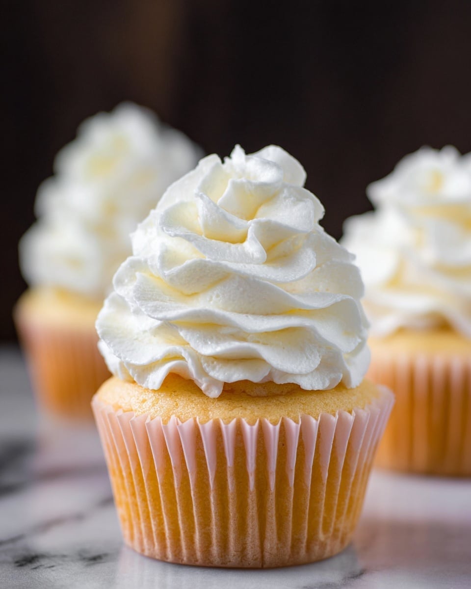 This close-up image shows a thick, creamy white frosting smoothly spread on a frosting spatula. The frosting is soft with a light, airy texture that has fine ridges and peaks formed by spreading. The background is softly blurred with a white marbled texture visible behind and below, hinting at a mixing bowl filled with more frosting. Photo taken with an iphone --ar 4:5 --v 7