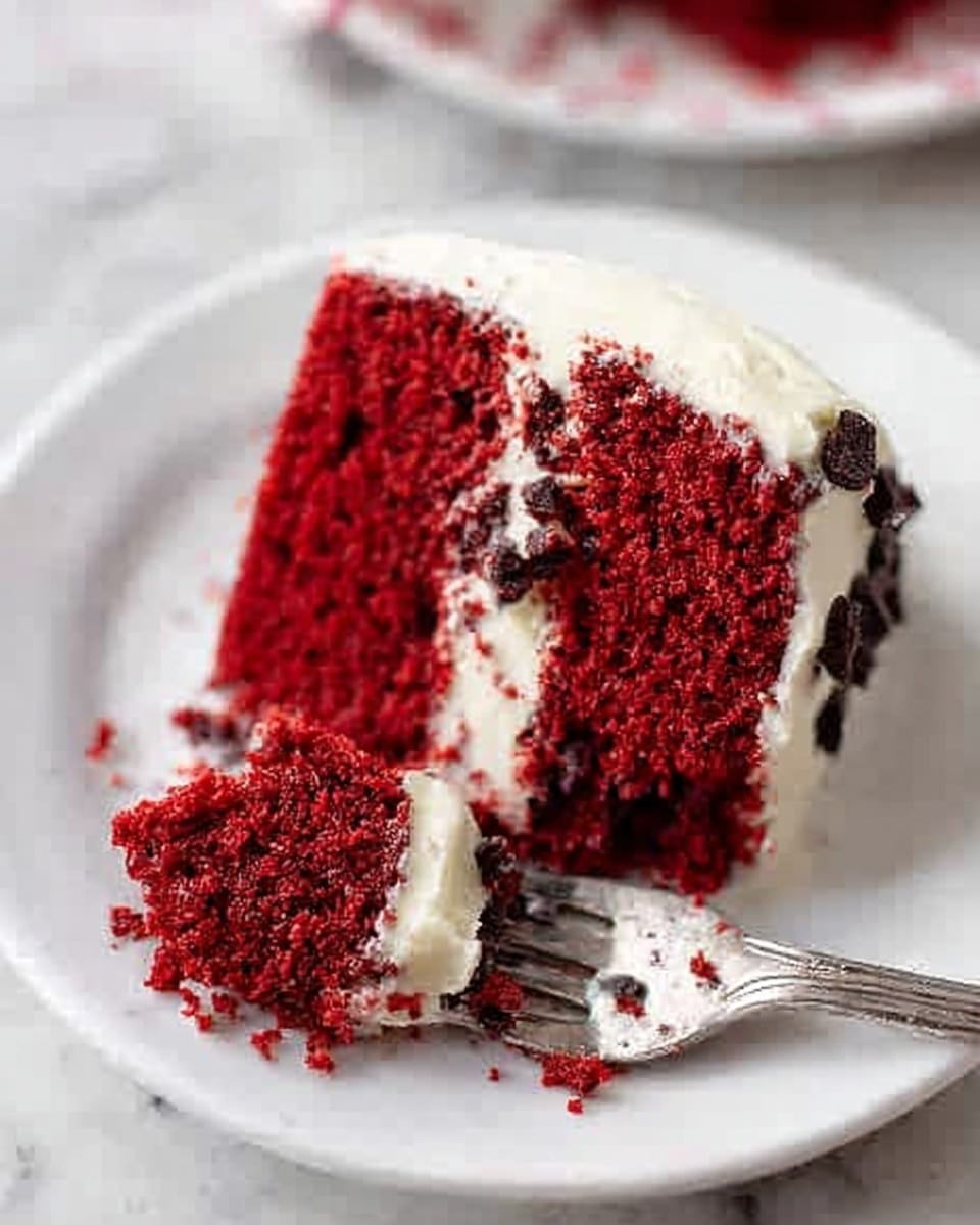 A close-up image of a single round slice of red velvet cake on a white plate placed on a white marbled surface. The cake has two layers: the bottom and top layers are bright red with a soft, moist texture speckled with dark chocolate chips. The cake is covered on the sides and top with a smooth, thick layer of creamy white frosting. A silver fork with a piece of cake on its tines rests on the plate next to the slice, with crumbs scattered around. A woman's hand is gently holding the fork lifting the bite. Photo taken with an iphone --ar 4:5 --v 7