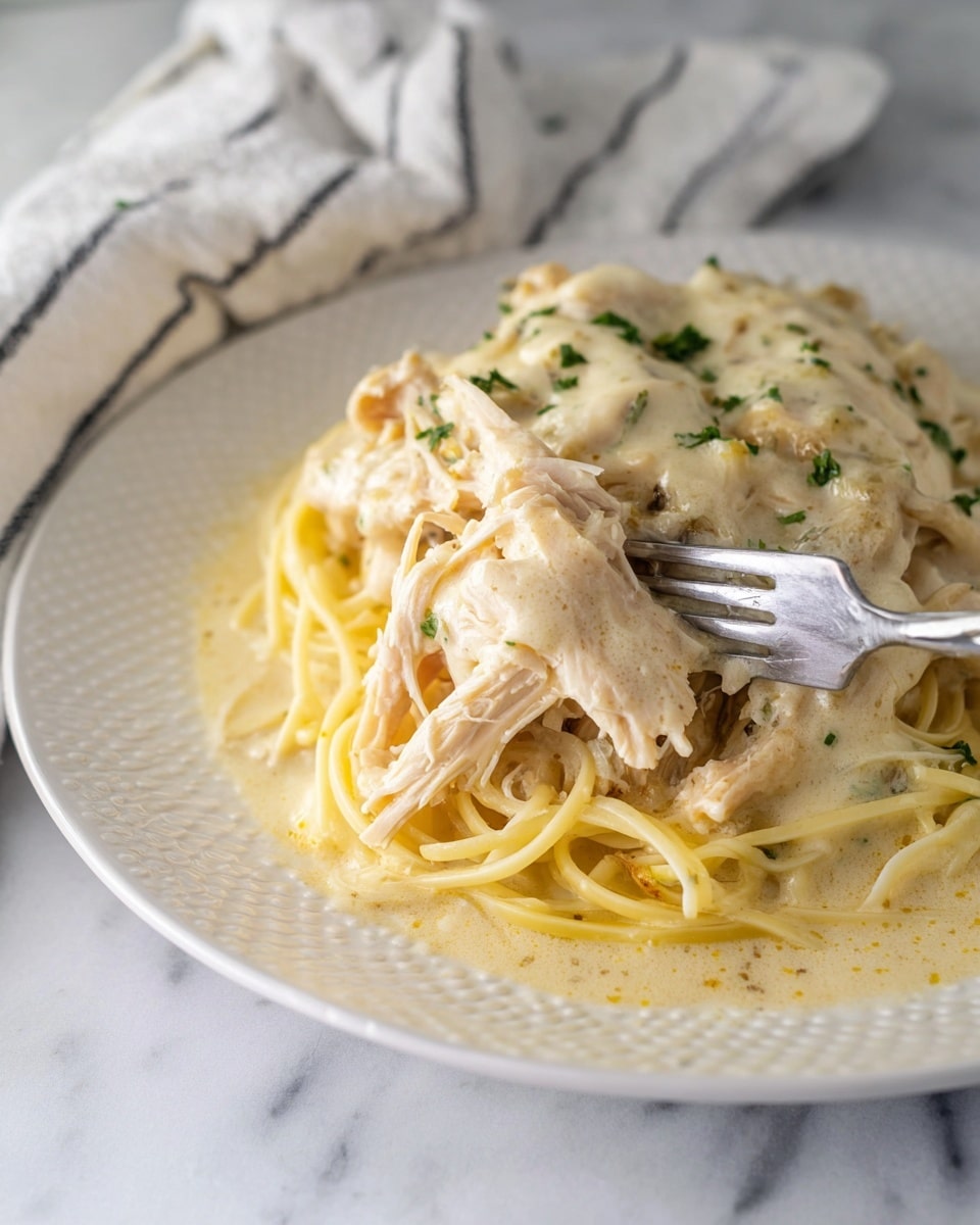 A white plate with a dotted texture holds two main parts: on the left is a thick filet of golden-brown chicken covered in a creamy white sauce sprinkled with green herbs, and on the right is a pile of white spaghetti noodles also coated in the same creamy sauce with visible garlic pieces and green herb flakes. A silver fork rests on the right edge of the plate, and a white cloth with black stripes lies underneath on a white marbled surface. photo taken with an iphone --ar 4:5 --v 7