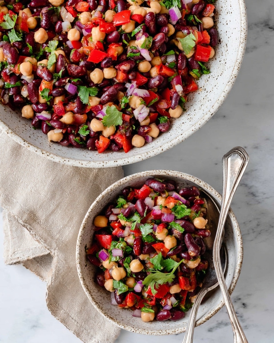 The image shows a colorful bean salad served in two white speckled bowls, one large and one smaller. The salad has three clear layers: the base layer consists of dark red kidney beans, black beans, and beige chickpeas, all mixed together with a matte texture. The second layer contains bright red chopped bell peppers and finely diced purple onions, adding fresh color and crunch. The top layer is scattered with small green leaves, likely cilantro or parsley, giving a fresh, leafy contrast. Two silver spoons rest inside the larger bowl on the right side. The bowls sit on a white marbled surface, with a beige cloth partially visible under the top bowl. photo taken with an iphone --ar 4:5 --v 7