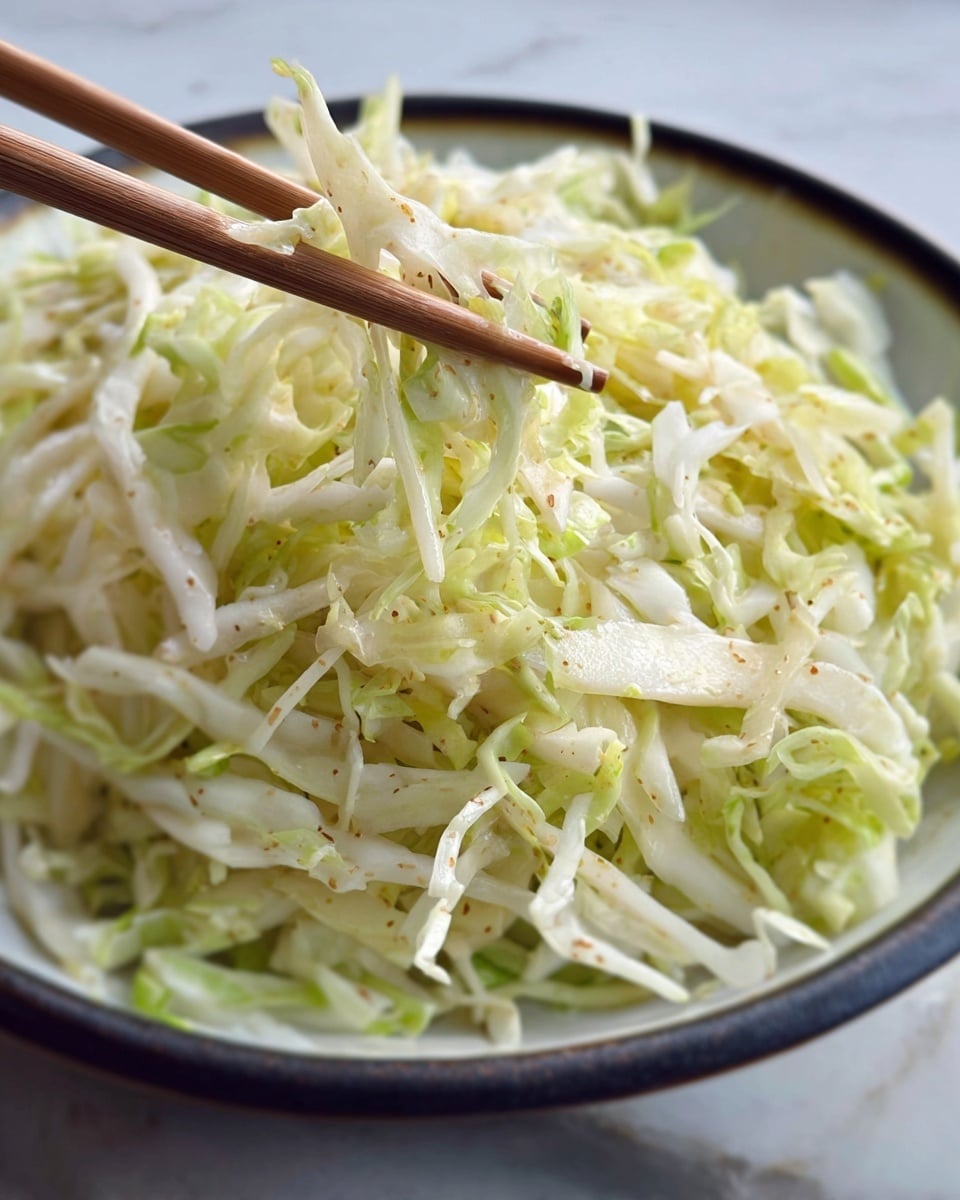 The image shows a white bowl filled with shredded light green and pale yellow cabbage piled high in one layer with a creamy white sauce underneath. The cabbage looks fresh and slightly moist, topped with a sprinkle of light brown crumbs or seasoning that adds texture and contrast. The bowl is placed on a white marbled surface with a soft cloth partially visible in the background. Photo taken with an iphone --ar 4:5 --v 7