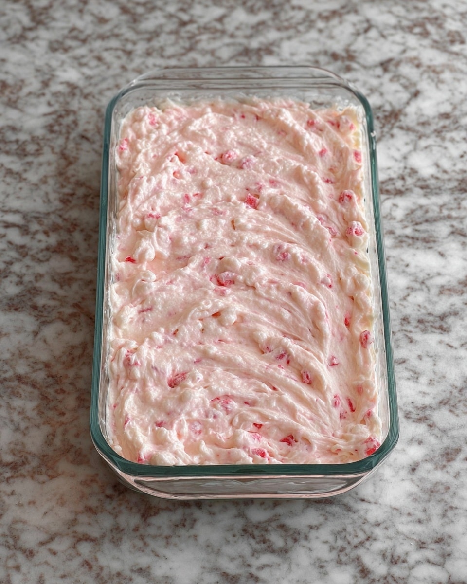 A clear glass rectangular dish filled with a thick, creamy mixture that is light pink in color with visible small chunks of red and white ingredients evenly spread throughout. The smooth top layer has gentle swirls and peaks, showing a soft and fluffy texture. The dish sits on a white marbled surface. photo taken with an iphone --ar 4:5 --v 7