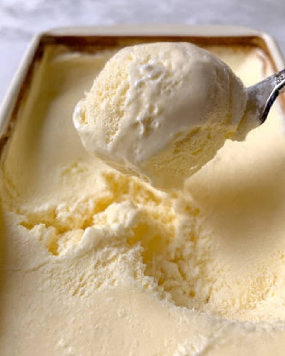 The image shows a close-up of a spoon scooping light yellow vanilla ice cream from a white dish. The ice cream looks creamy and smooth with a soft texture. The spoon is shiny silver and the scoop is slightly rounded with some ice cream dropping back into the dish. The background is a white marbled texture. Photo taken with an iphone --ar 4:5 --v 7