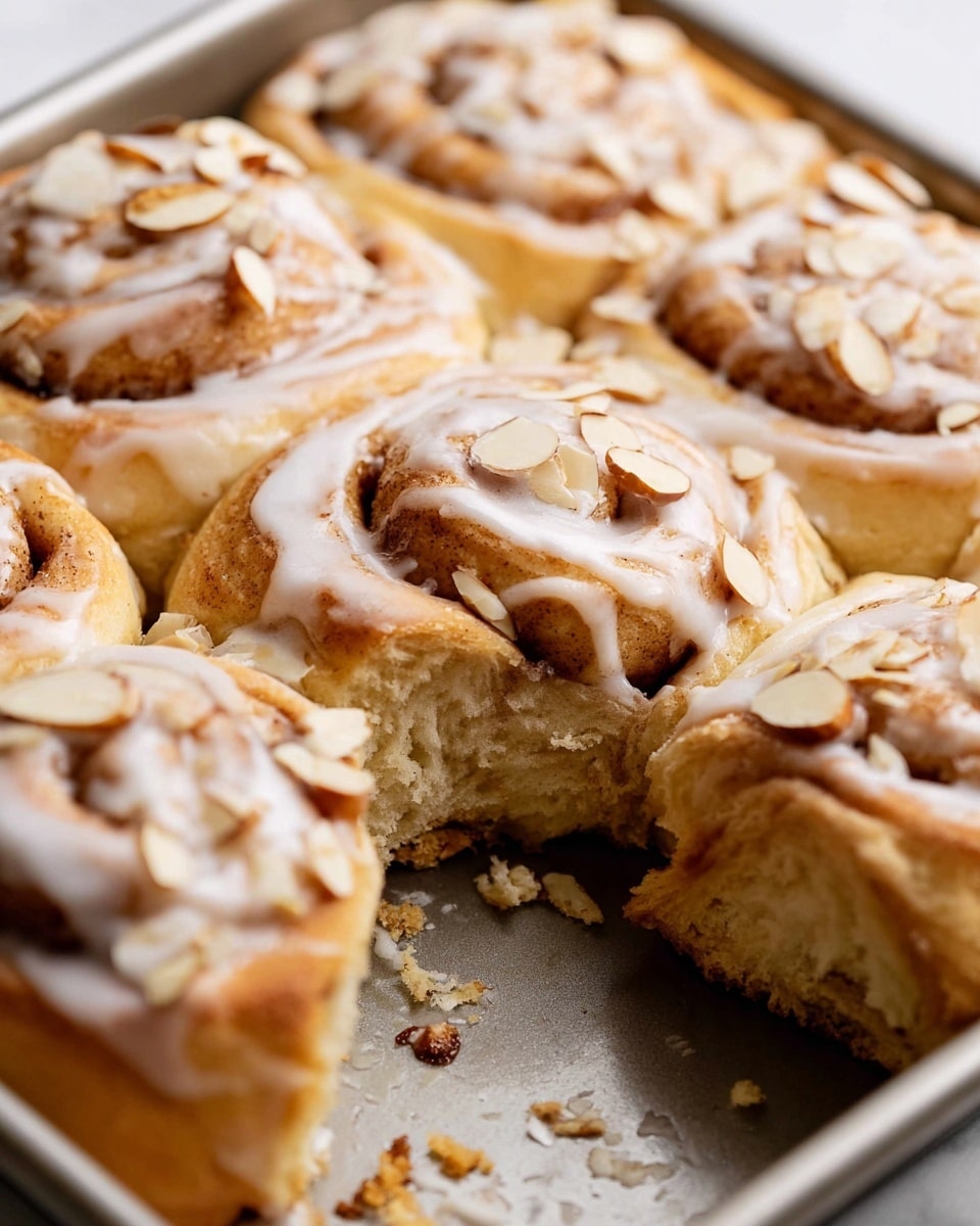 The dish shows a group of nine cinnamon rolls arranged closely together in a white pan, each roll with a golden-brown baked dough base that swirls inward with a cinnamon filling. A thick layer of creamy white icing is generously spread over the top, dripping down the sides and pooling slightly between the rolls. Thin slices of light beige almonds with darker edges are scattered over the icing, adding texture and contrast. The entire setup is placed on a white marbled surface. photo taken with an iphone --ar 4:5 --v 7
