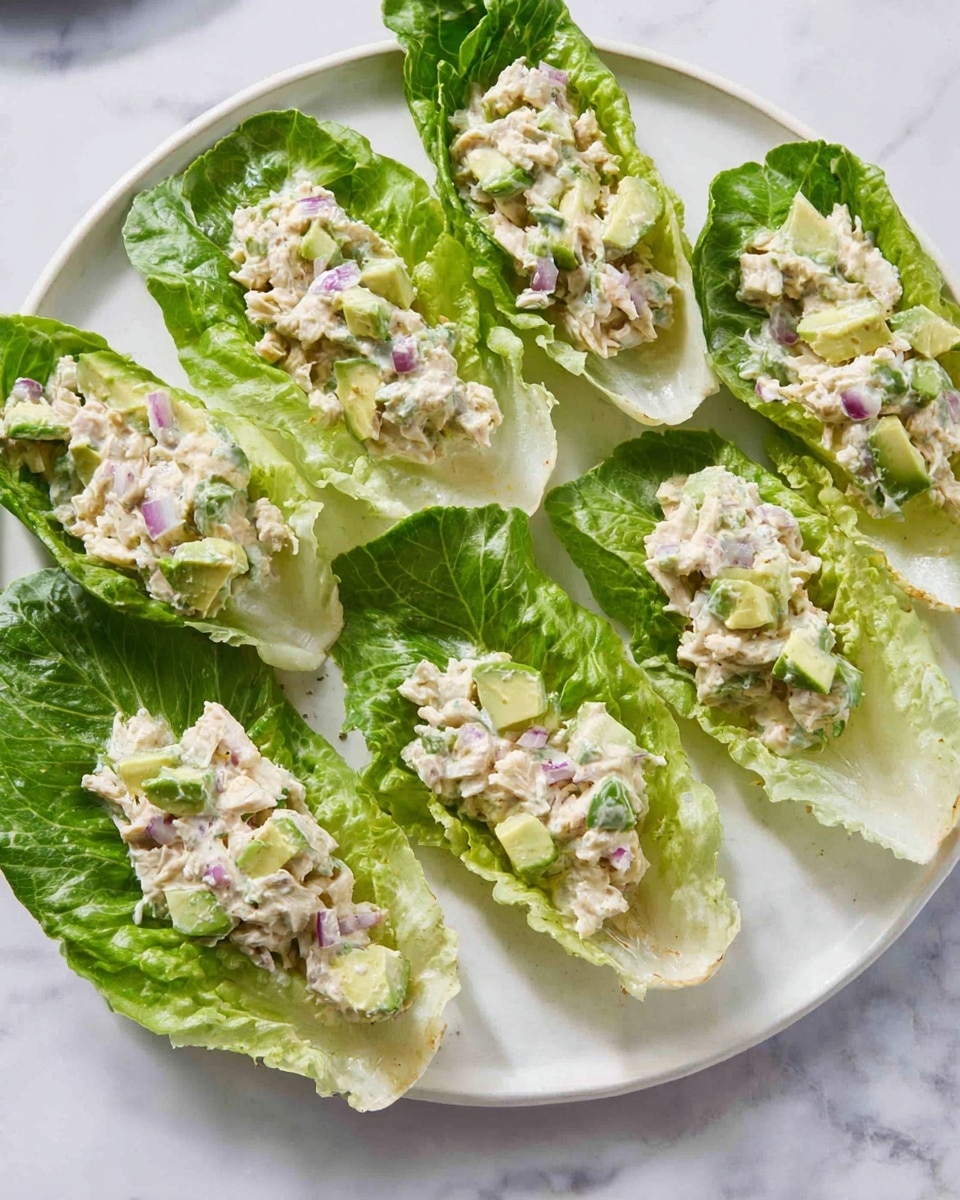 The image shows six fresh green romaine lettuce leaves arranged on a white plate with a white marbled texture surface, each leaf topped with a creamy tuna salad mix. The tuna salad has a light beige color with chunks of pale green avocado, small pieces of red onion, and some green herbs mixed in, giving it a soft, textured look. The lettuce leaves are curved, creating little bowls that hold the salad, and the overall presentation looks fresh and light. photo taken with an iphone --ar 4:5 --v 7