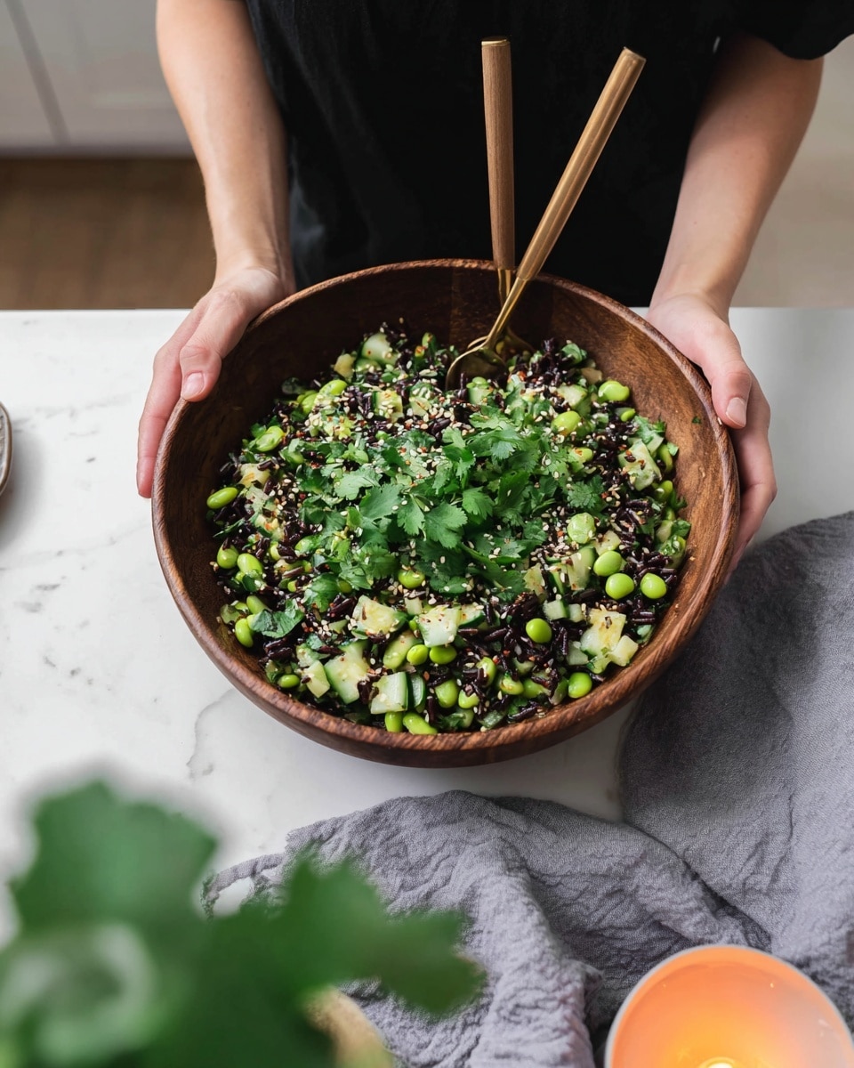 A dark brown bowl filled with a fresh salad sits on a white marbled surface, held by a person wearing a black shirt. The salad has three visible layers: a base of bright green edamame and zucchini pieces, mixed with small black rice grains, and topped with fresh green cilantro leaves and sprinkled white sesame seeds. Two wooden-handled forks rest inside the bowl. In the foreground, an orange candle and some blurred green leaves add warmth and depth to the scene. photo taken with an iphone --ar 4:5 --v 7