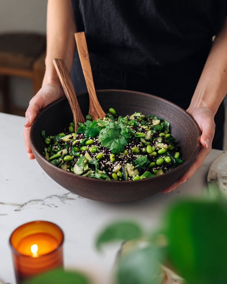 A large dark brown wooden bowl contains a mixed salad, held by a woman’s hands at both sides. The salad has three main layers: bright green edamame beans scattered throughout, diced light green cucumber pieces, and dark black rice or beans mixed in. On top, fresh bright green cilantro leaves are spread evenly with a sprinkle of white sesame seeds and small red pepper flakes for texture. Two wooden-handled metal spoons stand upright in the salad, placed near the center of the bowl. The bowl sits on a white marbled surface with a soft gray cloth on the right side and a blurry orange candle and green plant in the foreground. The person holding the bowl wears a black shirt. photo taken with an iphone --ar 4:5 --v 7
