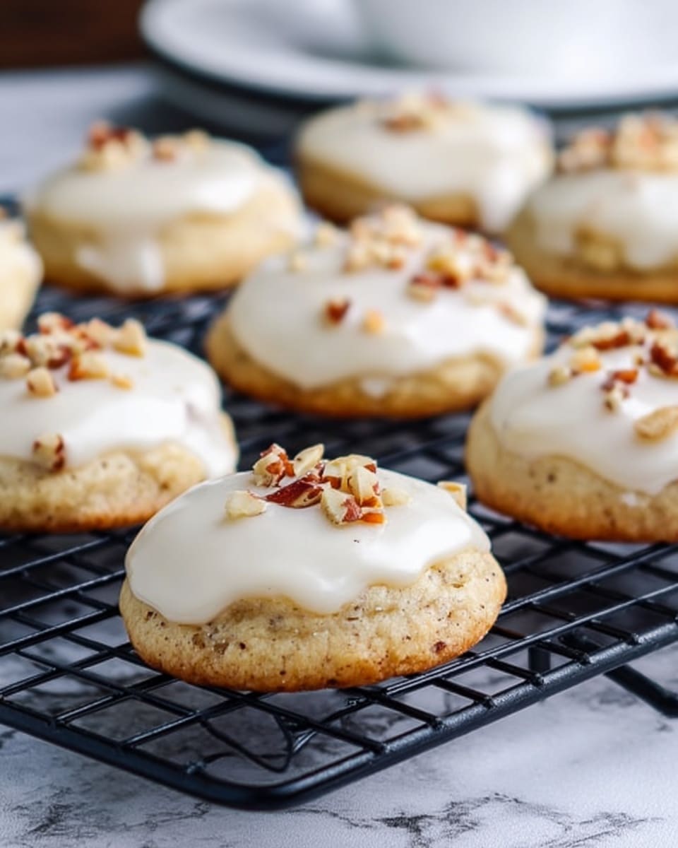 Small round iced cookies sit on a black cooling rack on top of a white marbled surface. Each cookie has two layers: a light golden-brown base with a slightly rough texture, topped with a thick, creamy white icing that looks smooth and glossy. On top of the icing, small pieces of chopped nuts add a crunchy texture and a touch of brown and tan color. In the background, a portion of a white plate is visible. photo taken with an iphone --ar 4:5 --v 7