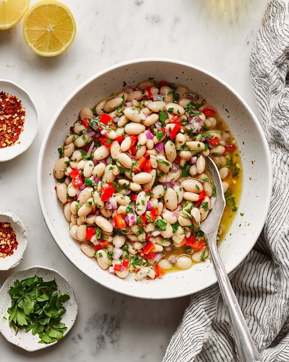 A white bowl filled with a colorful bean salad made of three main layers: the base layer of creamy white beans, mixed with small pieces of bright red bell peppers and finely chopped green herbs, and tiny bits of pale purple red onion throughout. The salad is tossed in a light golden oil, seen pooling slightly in the middle. A silver spoon is resting inside the bowl, with its handle stretching out over the edge. The bowl sits on a white marbled surface, surrounded by small white dishes holding red pepper flakes and fresh green herbs, along with a half lemon at the top left and a striped cloth napkin at the bottom right. photo taken with an iphone --ar 4:5 --v 7