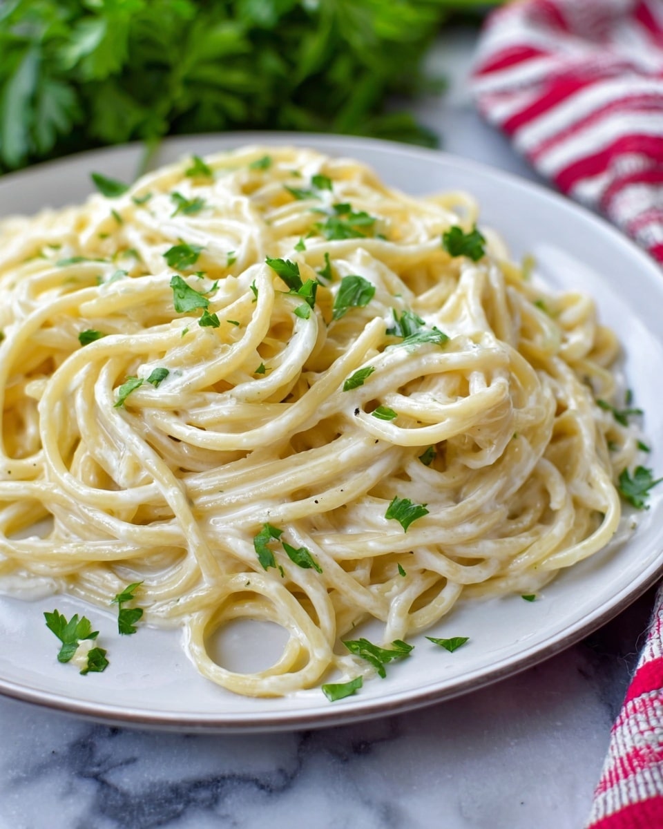 A close-up view of creamy spaghetti pasta piled on a white plate, with smooth, slightly shiny, pale yellow noodles coated evenly in a thick white sauce. Small green parsley leaves are scattered on top, adding a touch of fresh color. The plate sits on a white marbled surface with a blurred background featuring a red and white striped cloth and green leafy herbs. Photo taken with an iphone --ar 4:5 --v 7