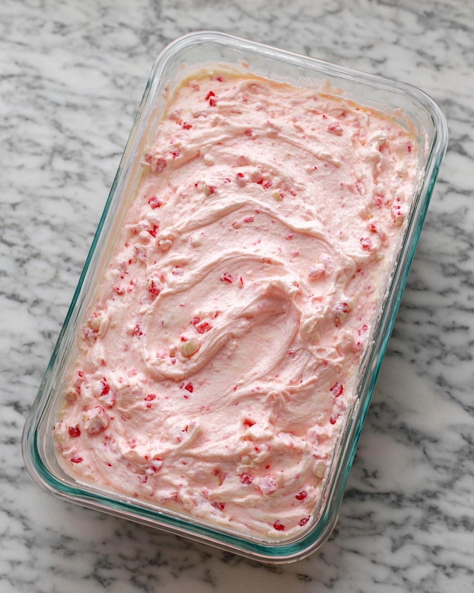 A rectangular clear glass dish is filled with a creamy, fluffy mixture that is pale pink with small bits of red and white scattered throughout. The surface of the mixture has a swirled texture, showing soft peaks and waves. The dish is placed on a white marbled textured surface. photo taken with an iphone --ar 4:5 --v 7