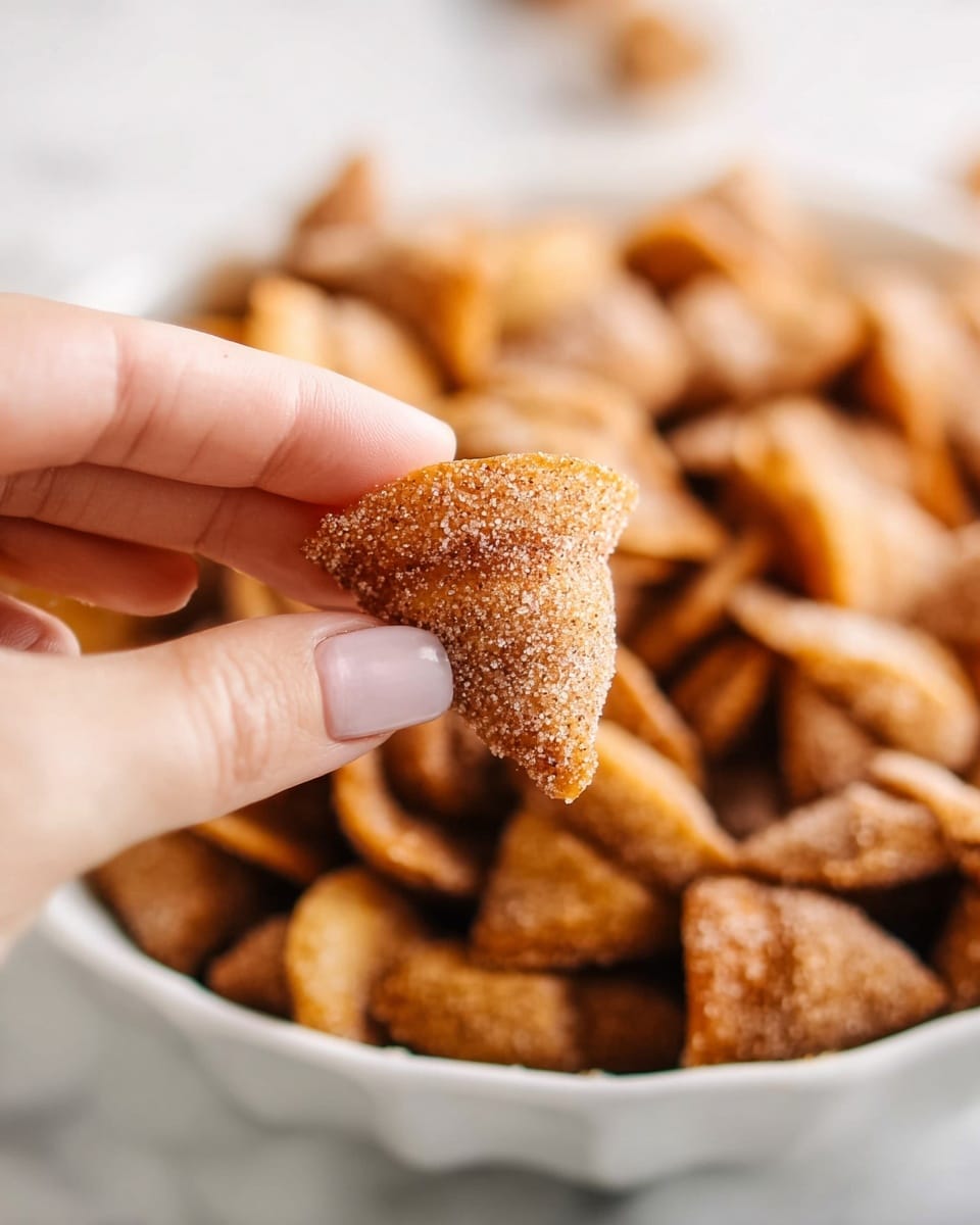 A close-up of a woman's hand holding a small, cone-shaped snack covered with sugar and cinnamon, with a textured, crispy surface. In the background, a large white bowl filled with many similar golden-brown, crunchy cone-shaped snacks coated in sugar and cinnamon is slightly out of focus. The scene is set against a white marbled surface. photo taken with an iphone --ar 4:5 --v 7