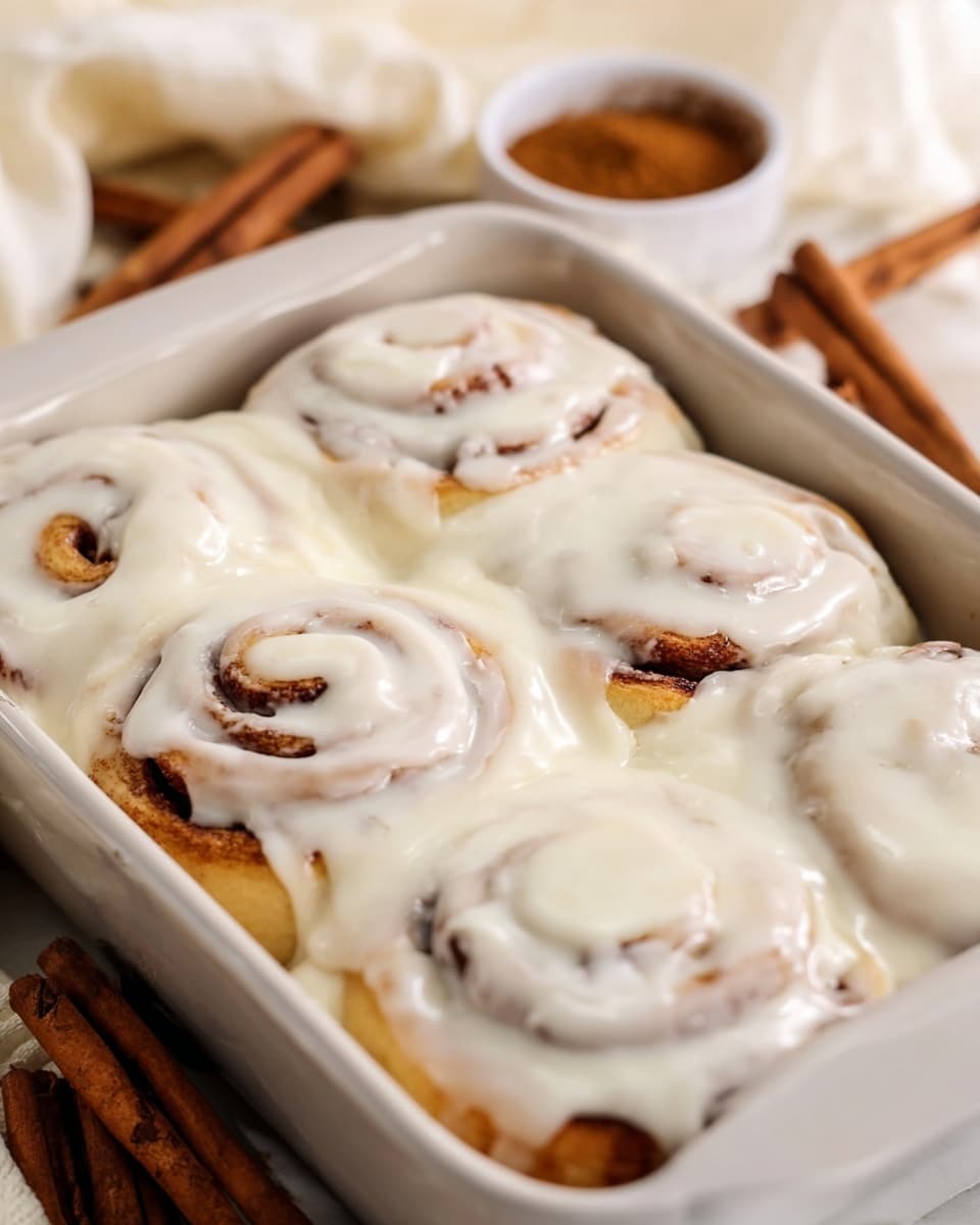 A close-up view of a white rectangular baking dish filled with six cinnamon rolls covered in a thick layer of creamy white icing that drips slightly over the edges. The cinnamon rolls have a golden brown swirl visible beneath the smooth, shiny icing that gives a soft texture look. The dish is placed on a light surface with cinnamon sticks and a small bowl of cinnamon powder blurred softly in the background, all on a white marbled texture. photo taken with an iphone --ar 4:5 --v 7