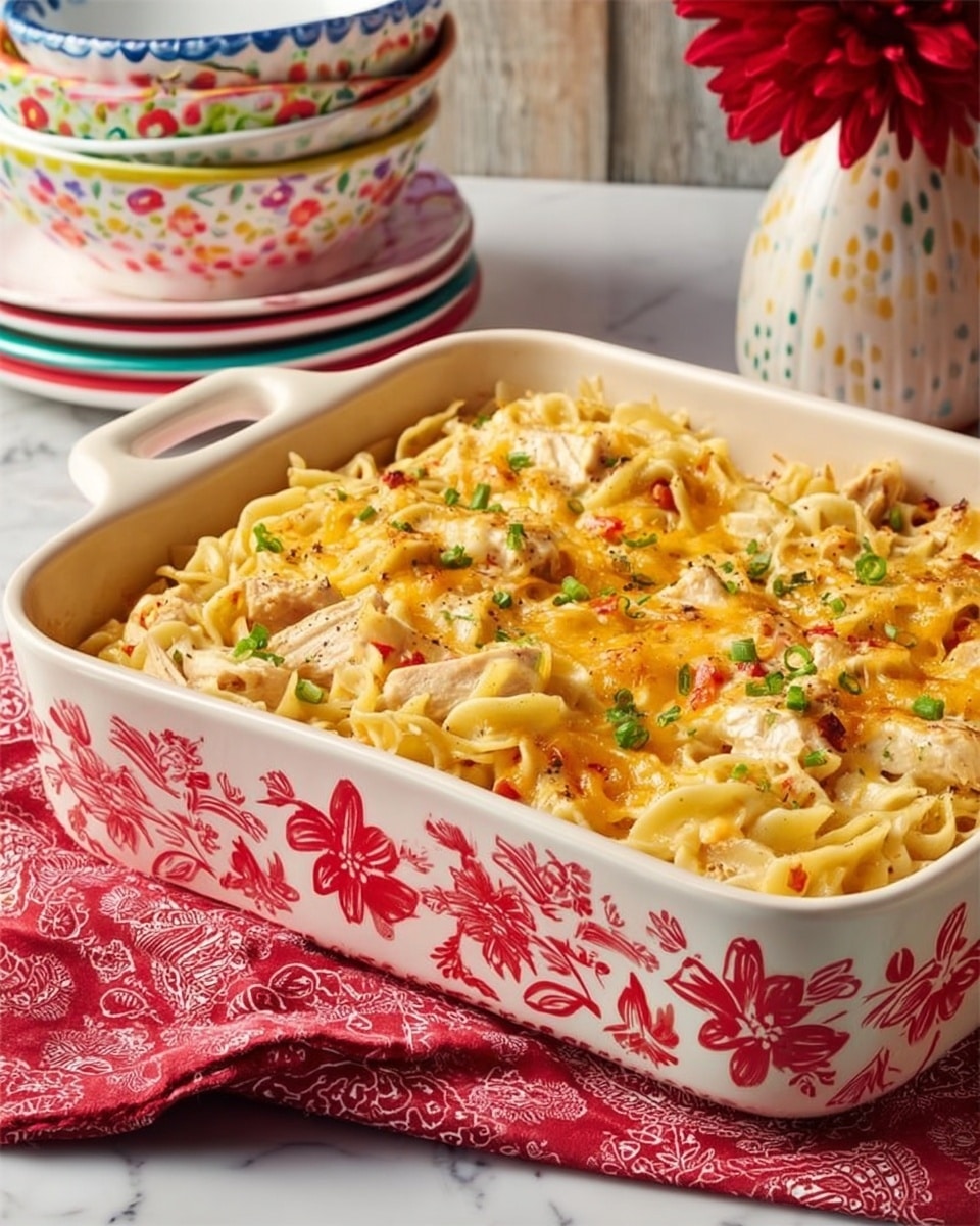 A white rectangular baking dish with a red floral pattern on the outside holds a baked noodle casserole. The casserole has one visible layer of creamy, cheesy noodles mixed with chunks of chicken and small bits of green peppers and red spices, creating a textured surface with shades of pale yellow, light brown, and hints of green and red. The noodles are lightly browned on top, giving a slightly crispy appearance. The dish is placed on a white marbled surface with a folded red patterned cloth underneath. In the background, there are stacked white bowls with colorful floral and dotted designs and a vase with a red flower, adding a warm and cozy feel to the scene. Photo taken with an iphone --ar 4:5 --v 7